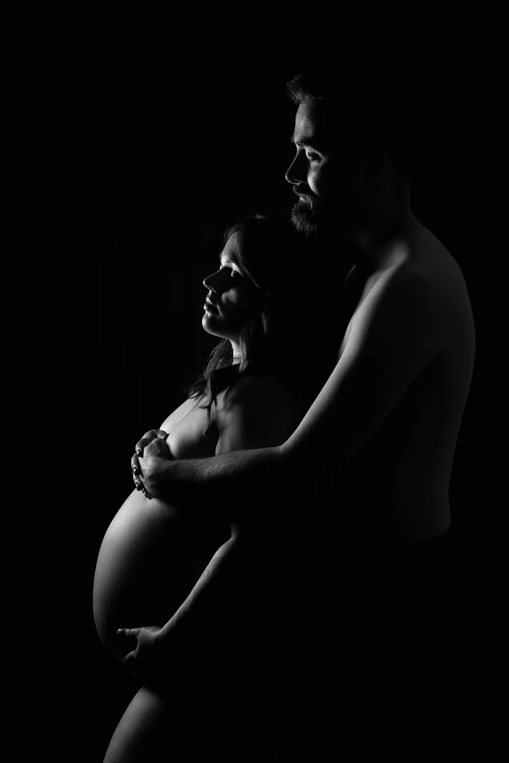 Couple en clair-obscur, mains sur le ventre, sur fond noir.