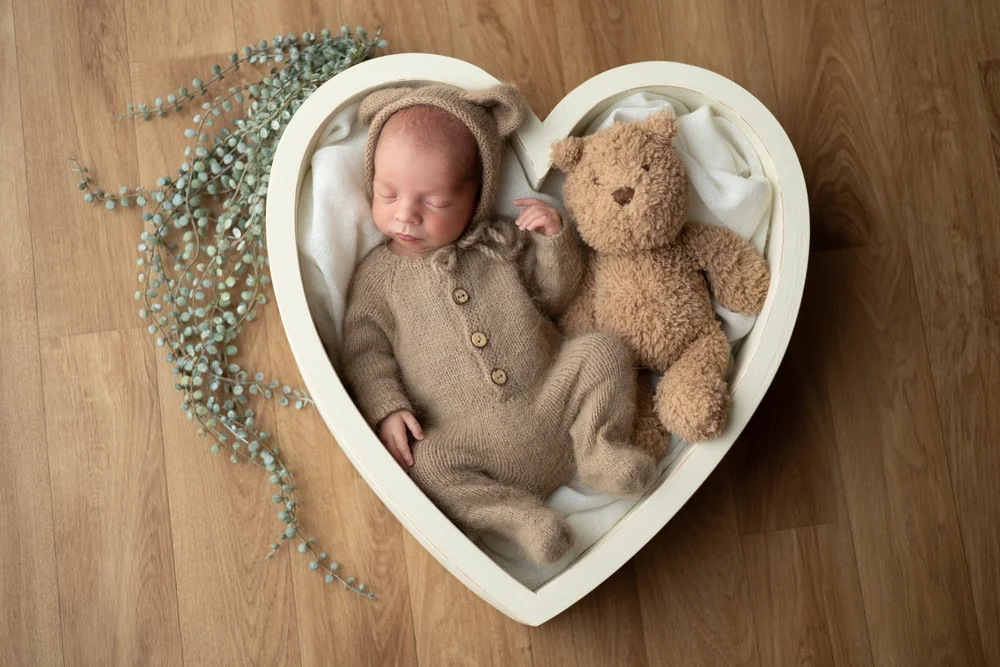 Nouveau-né en tenue marron avec un ours en peluche dans un panier en cœur.