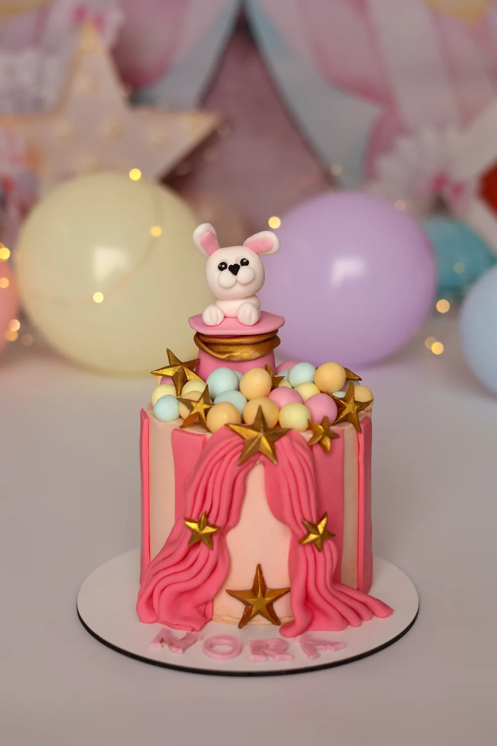 Gâteau d’anniversaire rose à rideaux devant des ballons pastel.