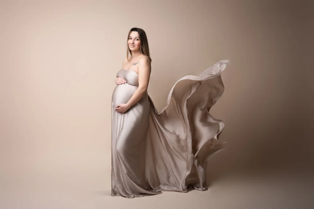Femme enceinte en robe grise dont le voile flotte, en studio sur fond beige.