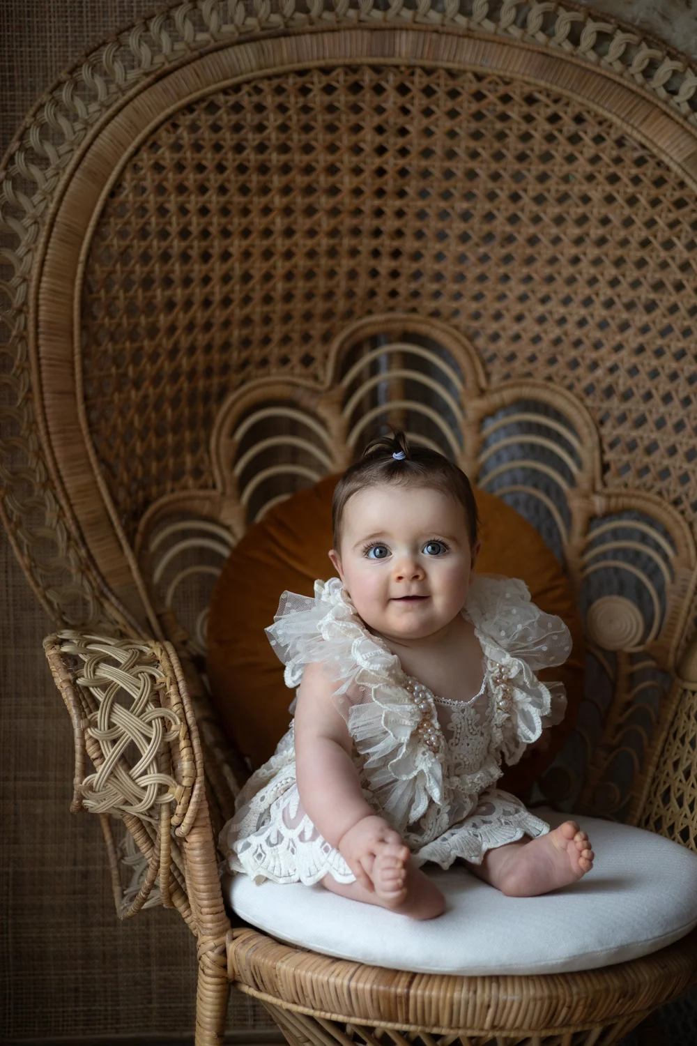 Bébé en robe de dentelle assise dans un grand fauteuil en rotin.
