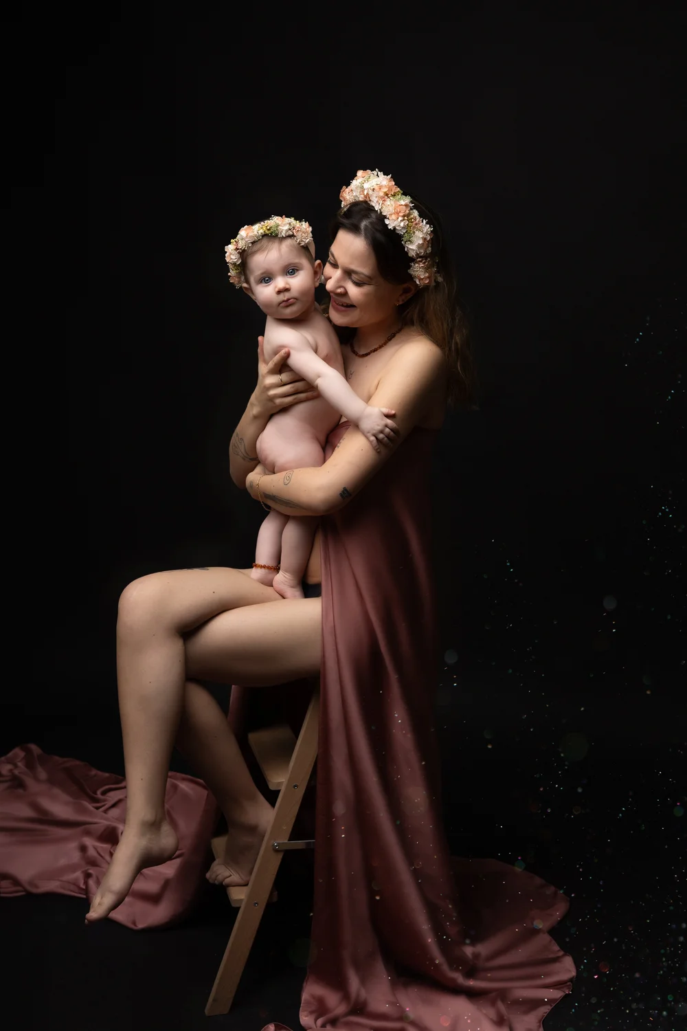 Maman en robe rose tenant son bébé, couronnes de fleurs, sur fond sombre.
