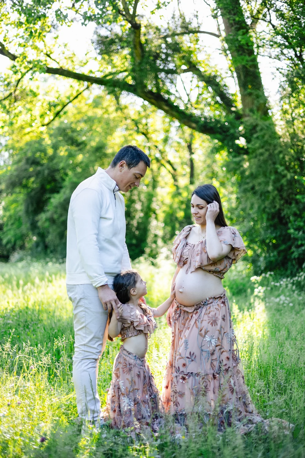 Futurs parents et leur fille en robes fleuries, dans un sous-bois ensoleillé.