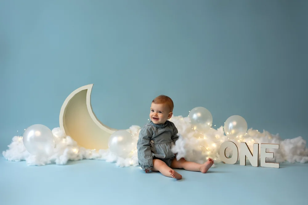Bébé assis devant un décor de lune et de ballons blancs, sur fond bleu.