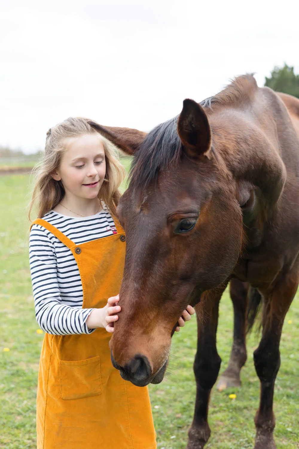 Fillette en salopette orange caressant un cheval dans un pré.