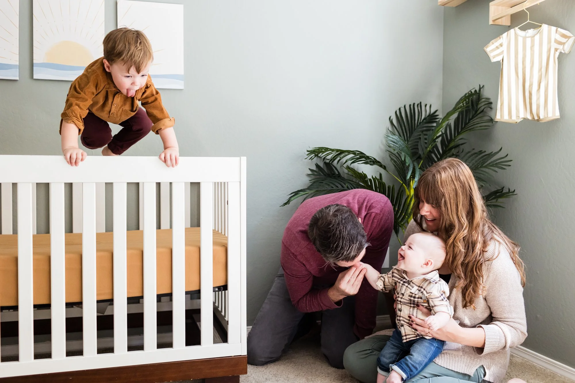 candid in-home family photography with toddler jumping in crib and parents laughing on ground with baby in Phoenix