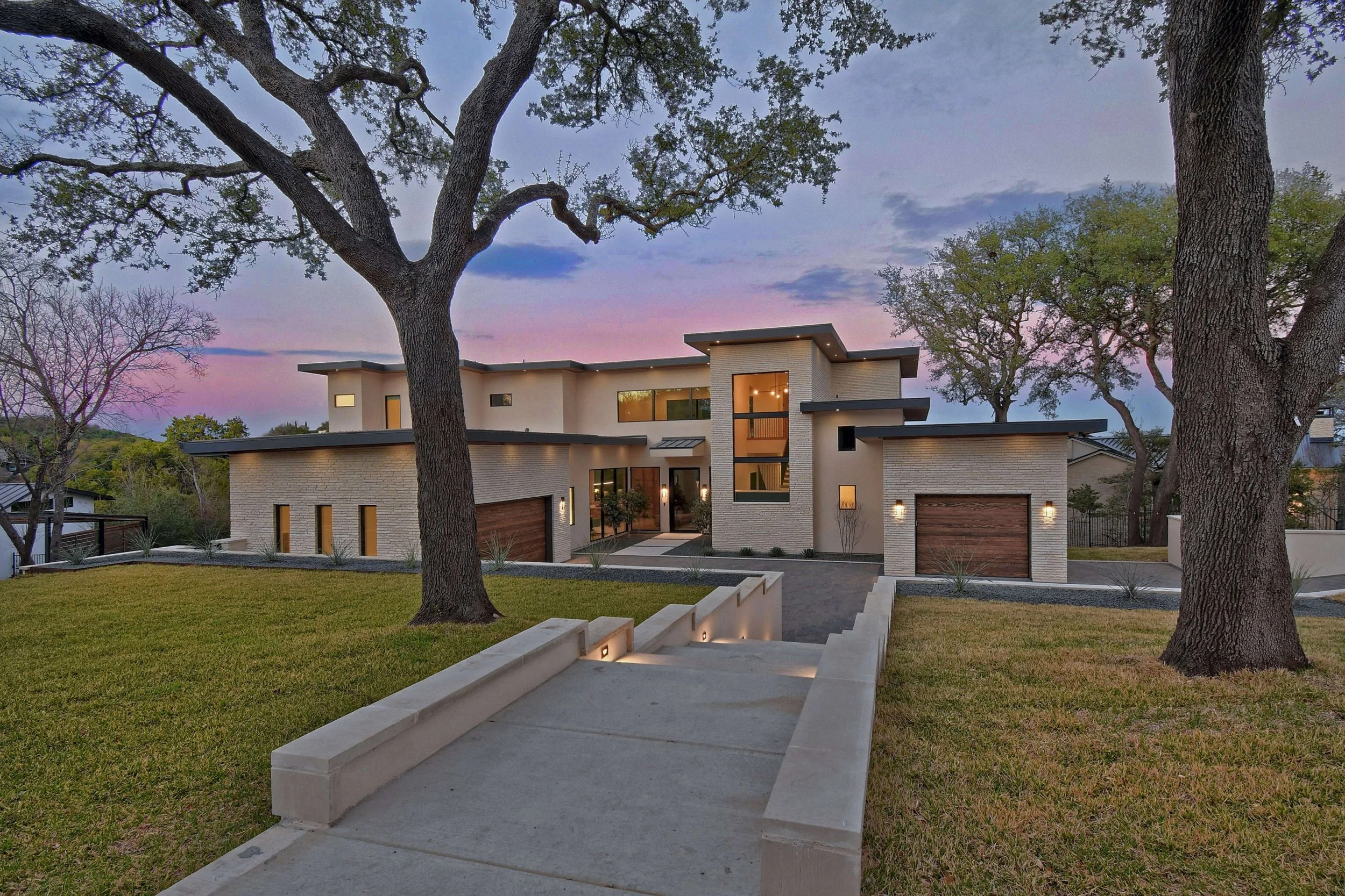 Modern two-story house with large windows, surrounded by trees and a lawn, during sunset.