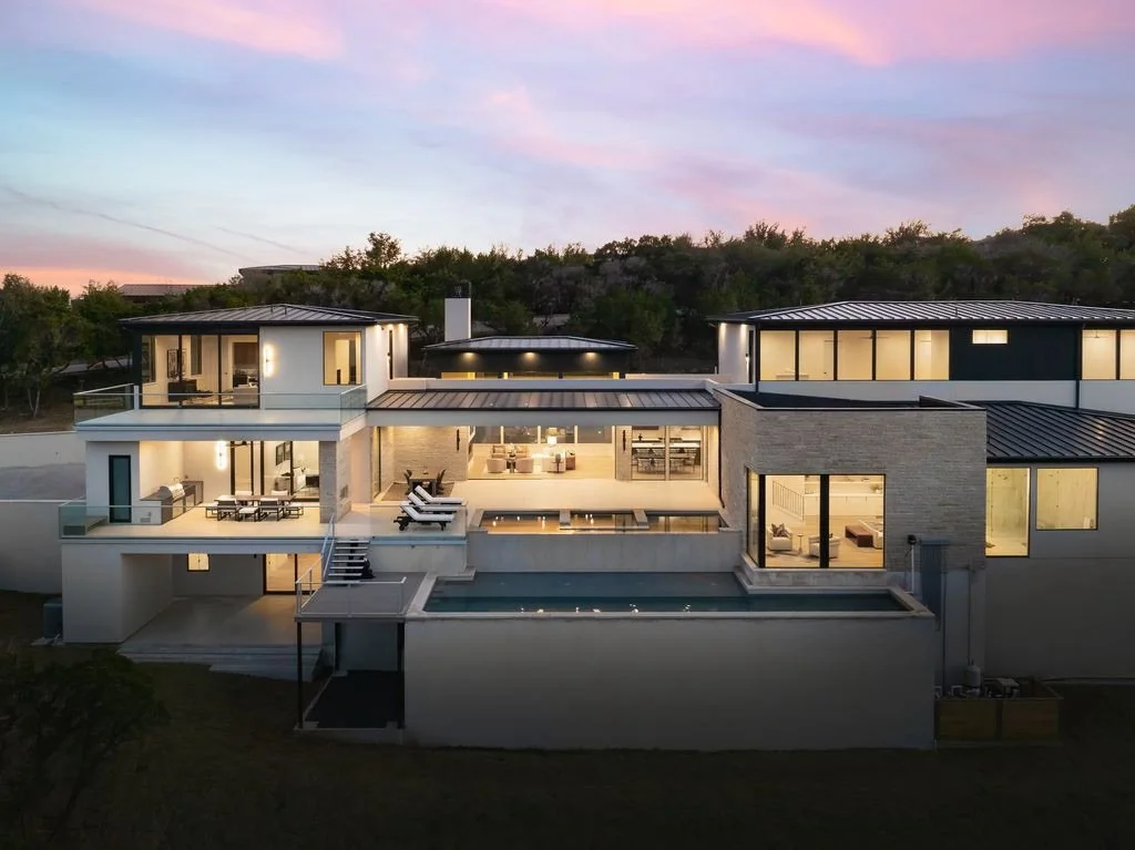 Modern multi-level house with large glass windows, outdoor swimming pool, and outdoor lounge areas, set against a sunset sky.