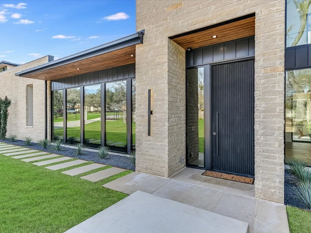 Modern house entrance with black door, large windows, wooden porch ceiling, and landscaped lawn.