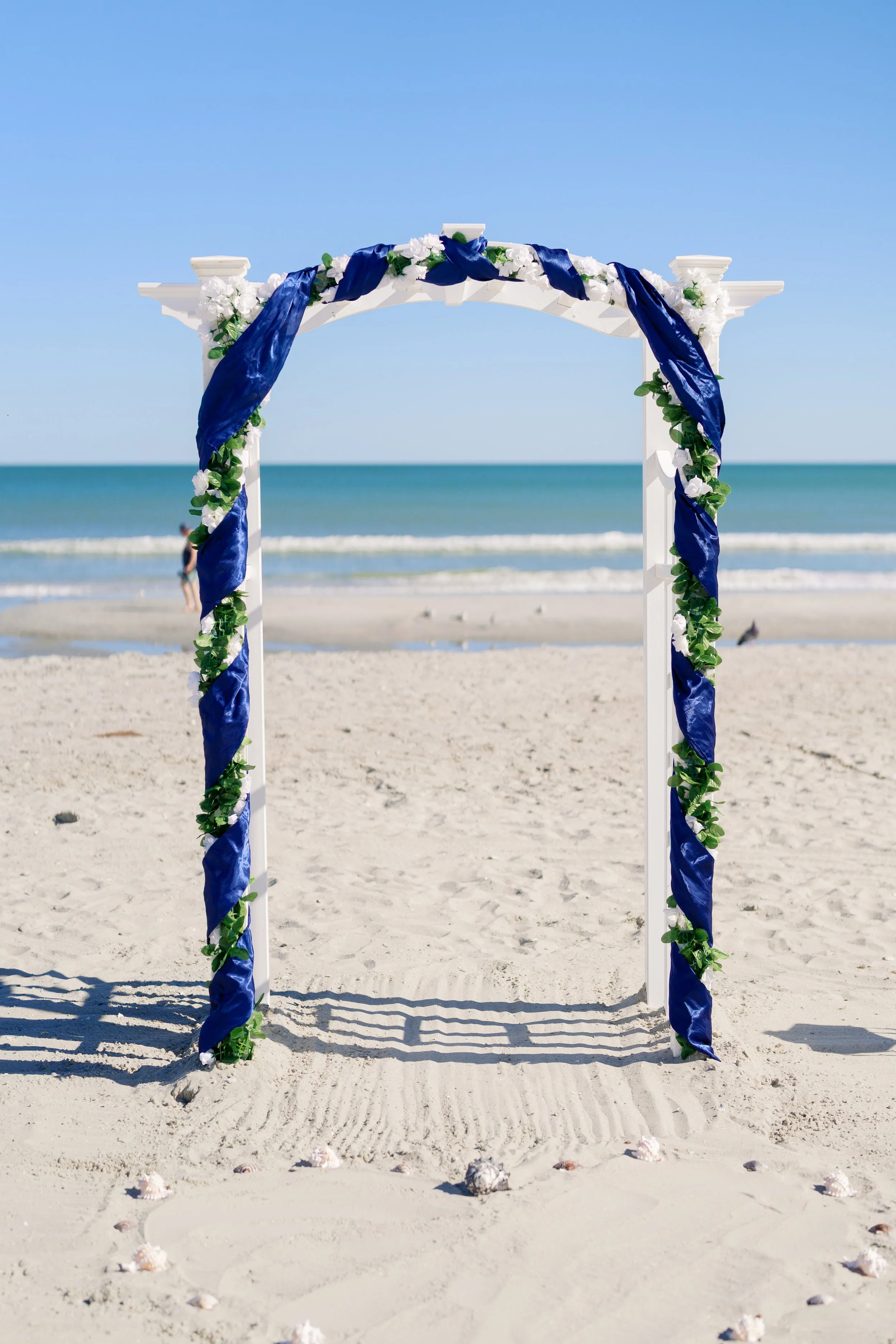 A wedding arch decorated with blue and white fabric and flowers on a sandy beach with ocean and sky in the background.