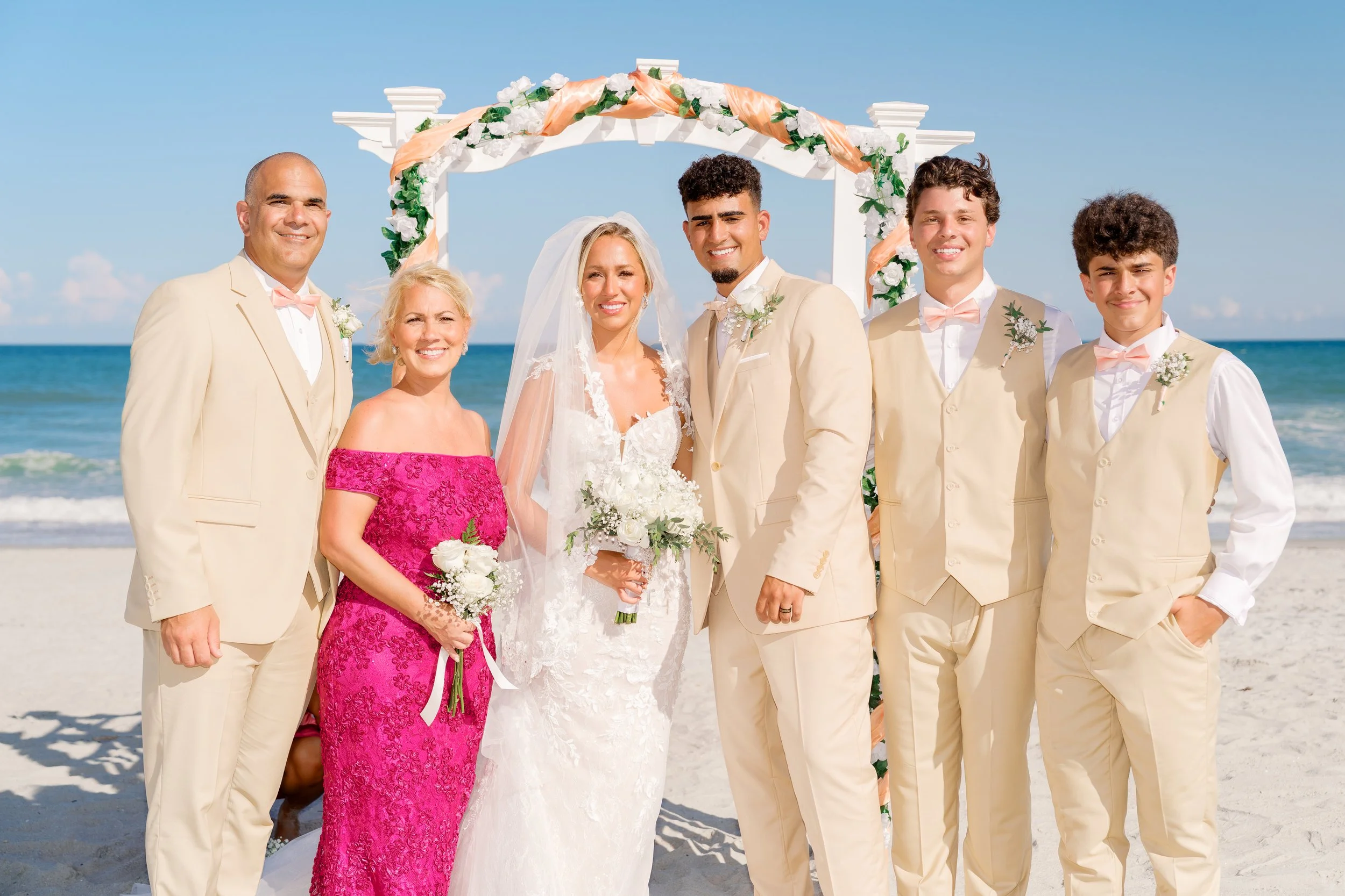A group of seven people dressed in wedding attire on the beach, standing under a decorated arch with white and peach accents, with the ocean and blue sky in the background.