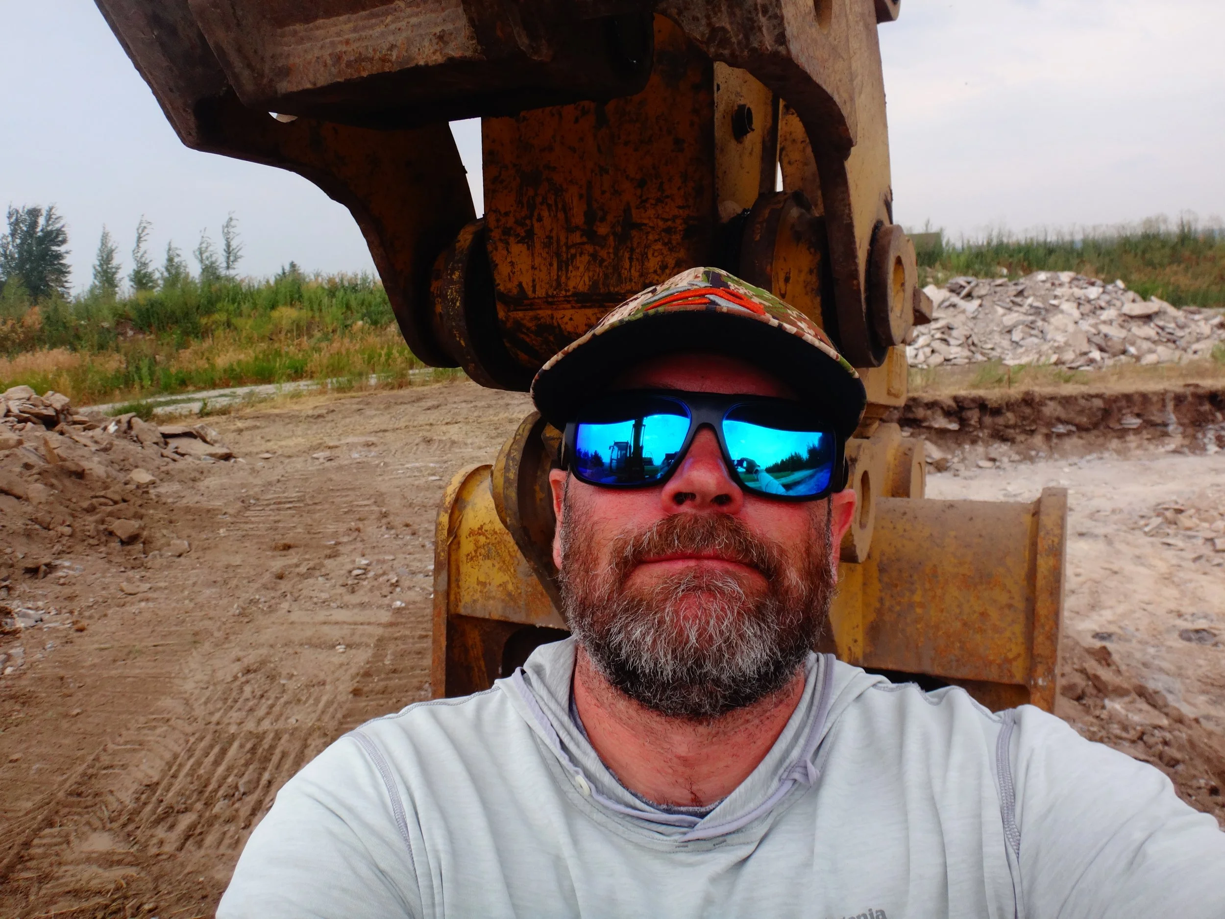 A man wearing sunglasses and a hat with a camouflage pattern taking a selfie in front of construction equipment at a construction site with dirt and trees in the background.
