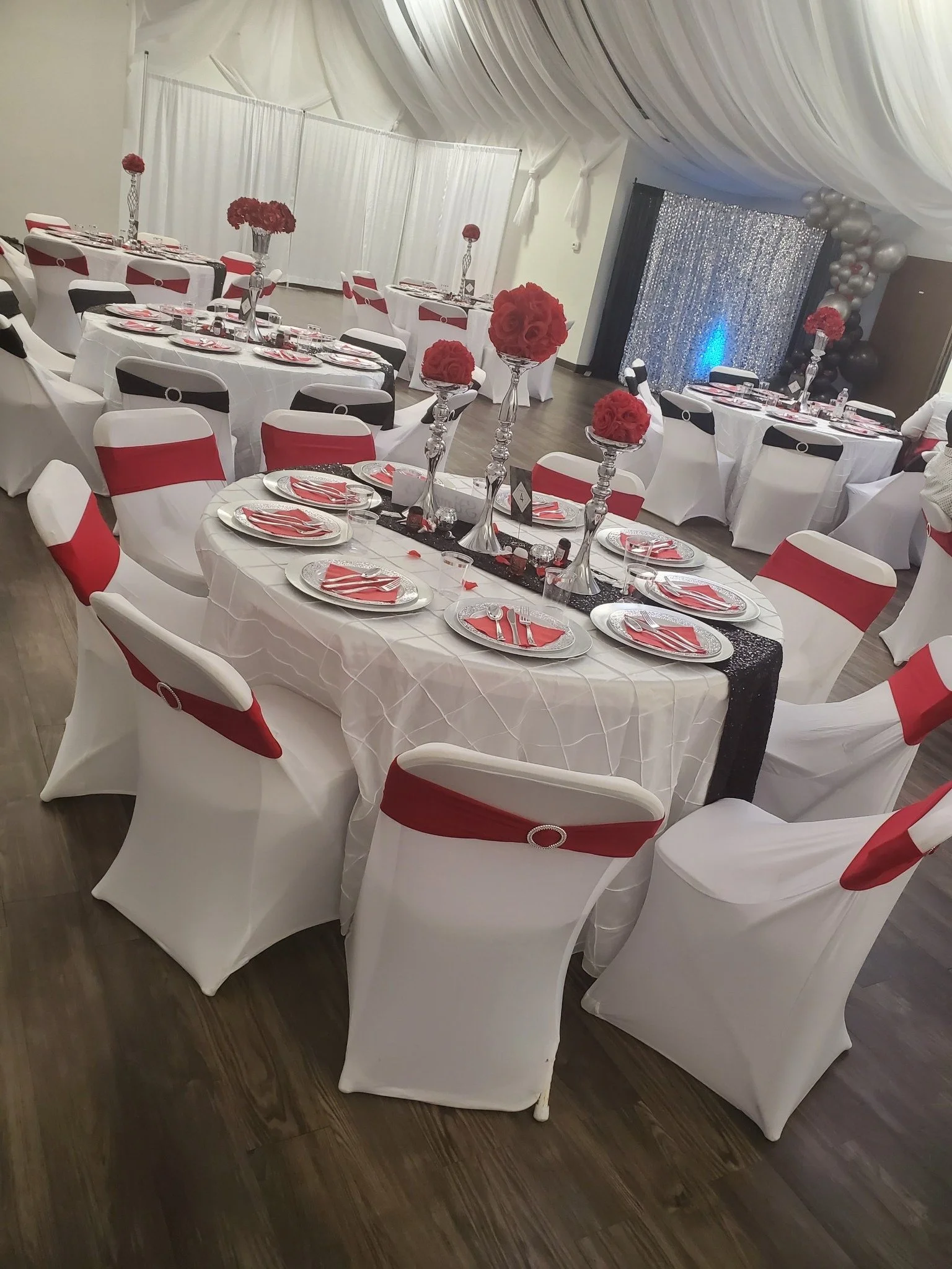 Wedding reception setup with white round tables, red and white chairs, and floral centerpieces featuring red flowers in tall silver vases. The backdrop has draped fabric and balloons.