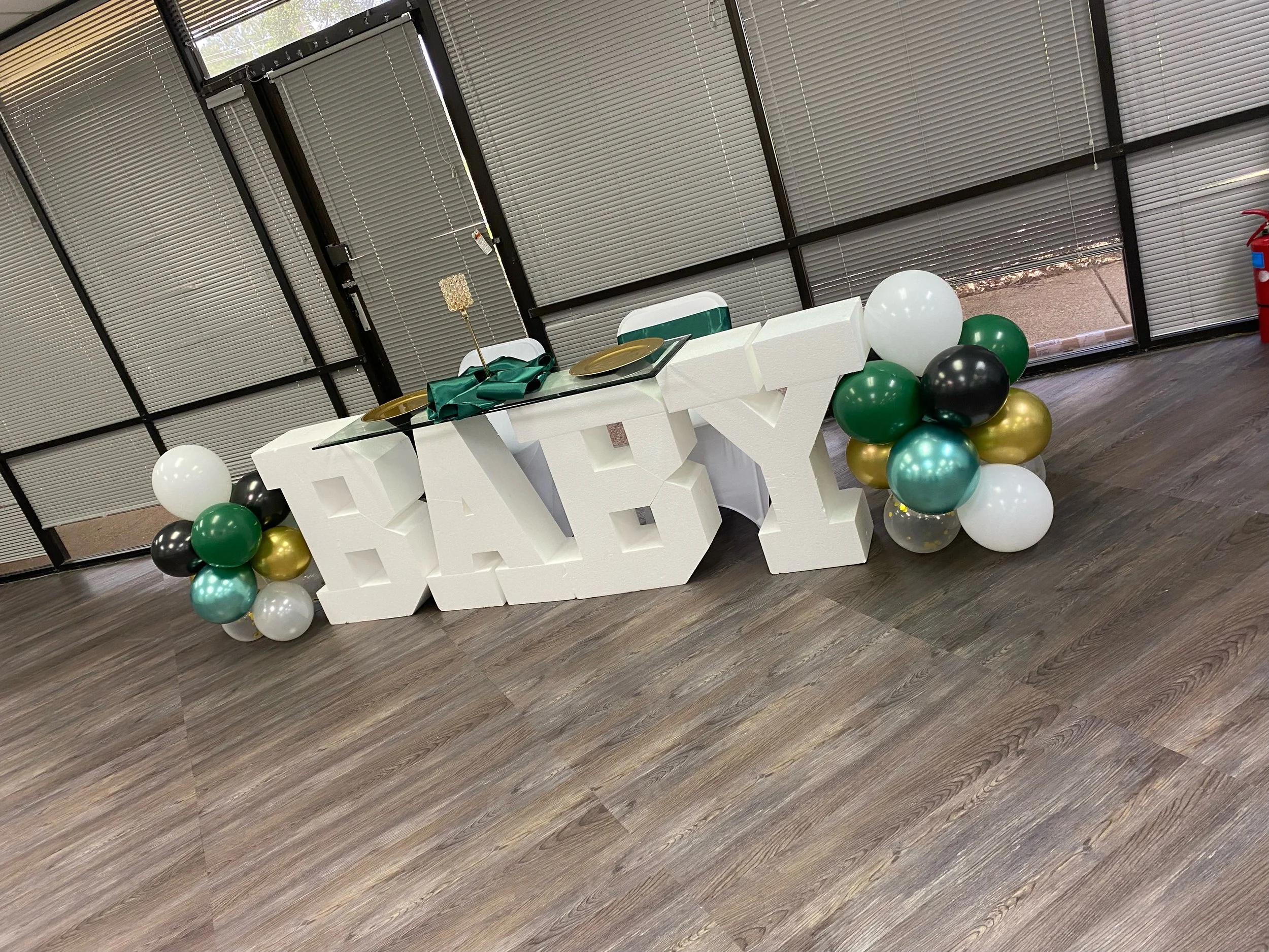 Balloon arrangement with gold, black, white, and green balloons, and decorative elements on a table, in a room with large window panels with closed blinds.