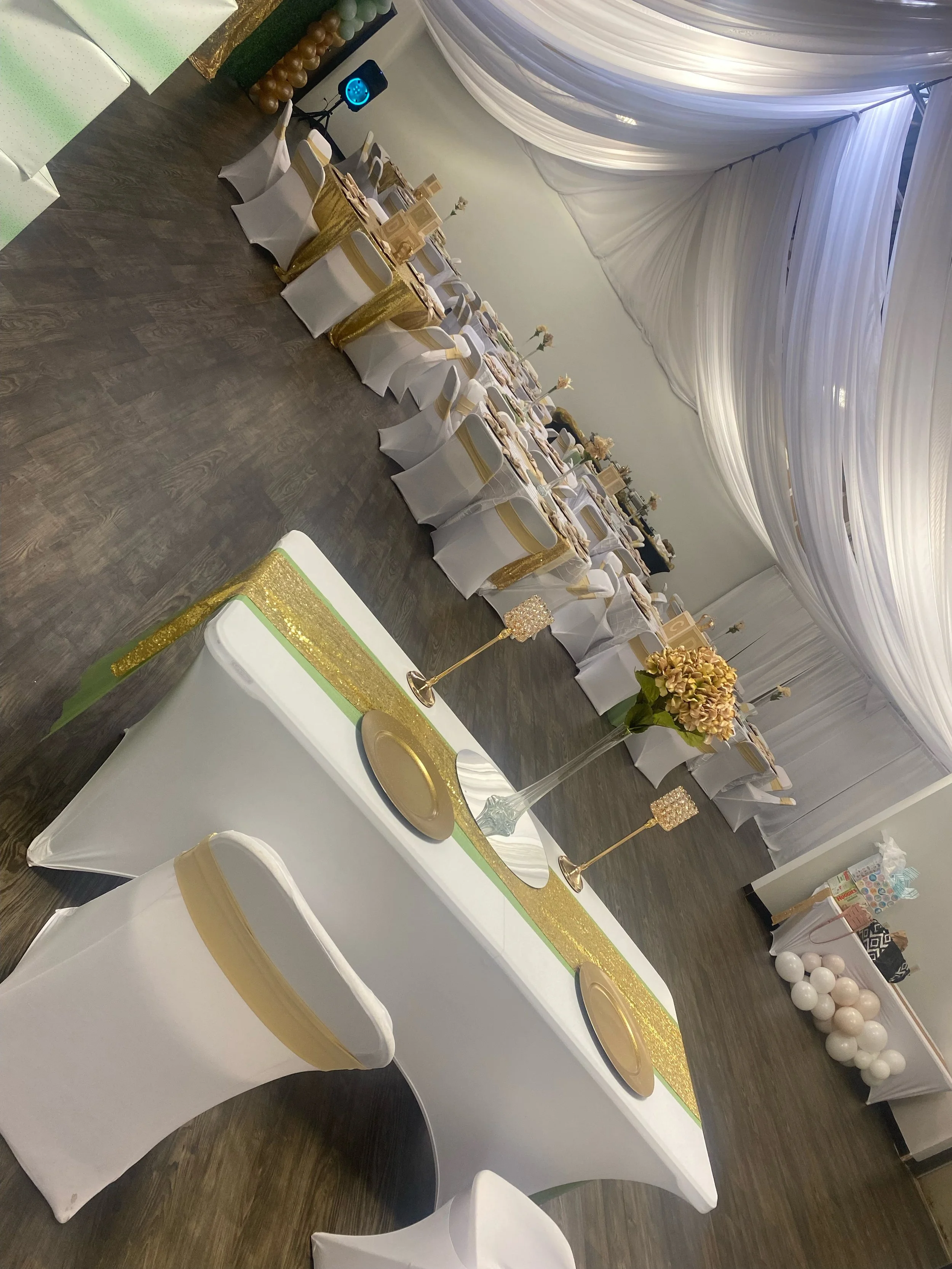 A decorated banquet hall with a long table set for a celebration, featuring white tablecloths, gold accents, floral centerpieces, and balloon decorations, with draped ceiling fabric in the background.