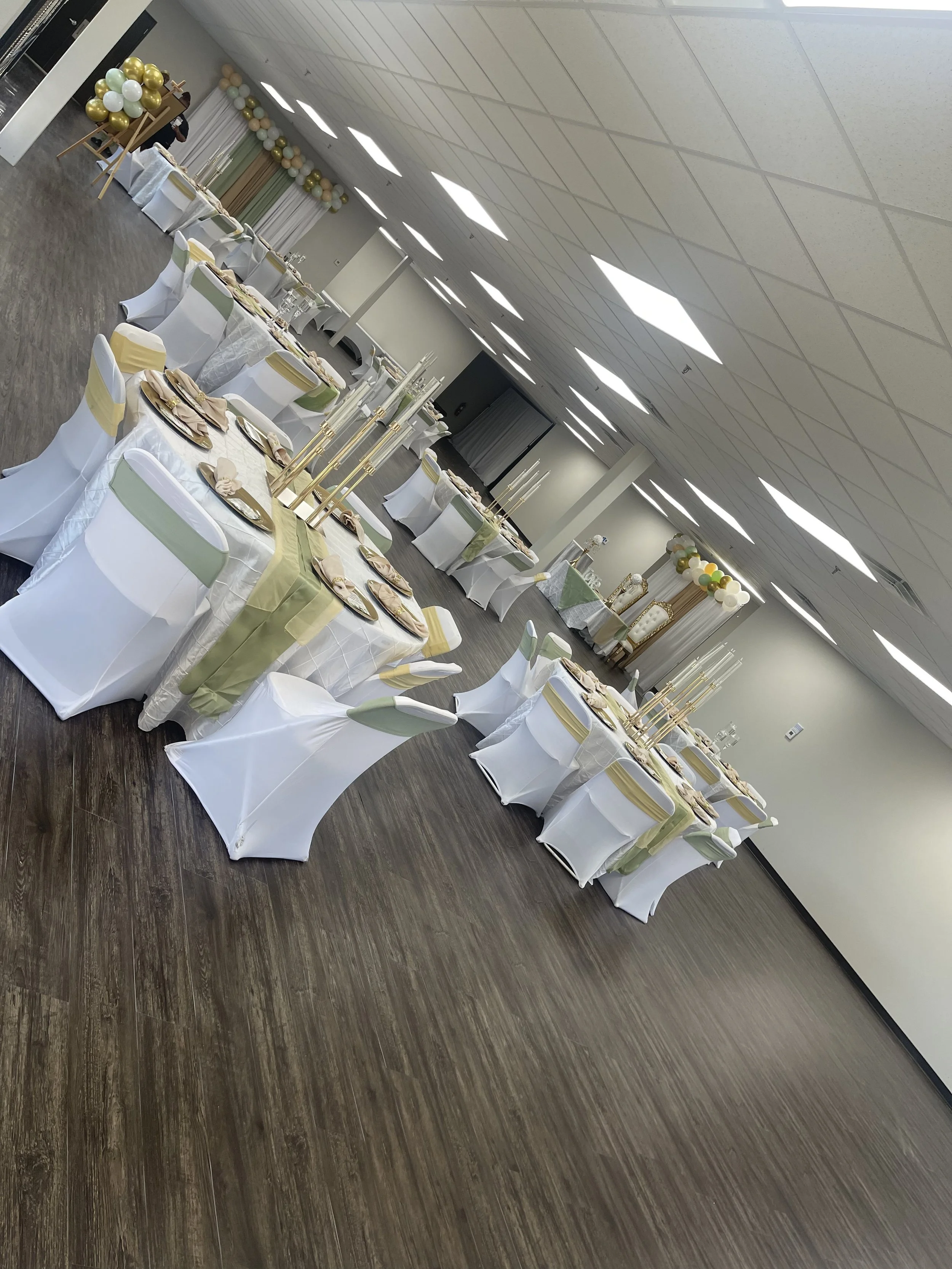 Event hall decorated with balloons, covered tables and chairs arranged for a celebration or reception, with a projector screen at the front.