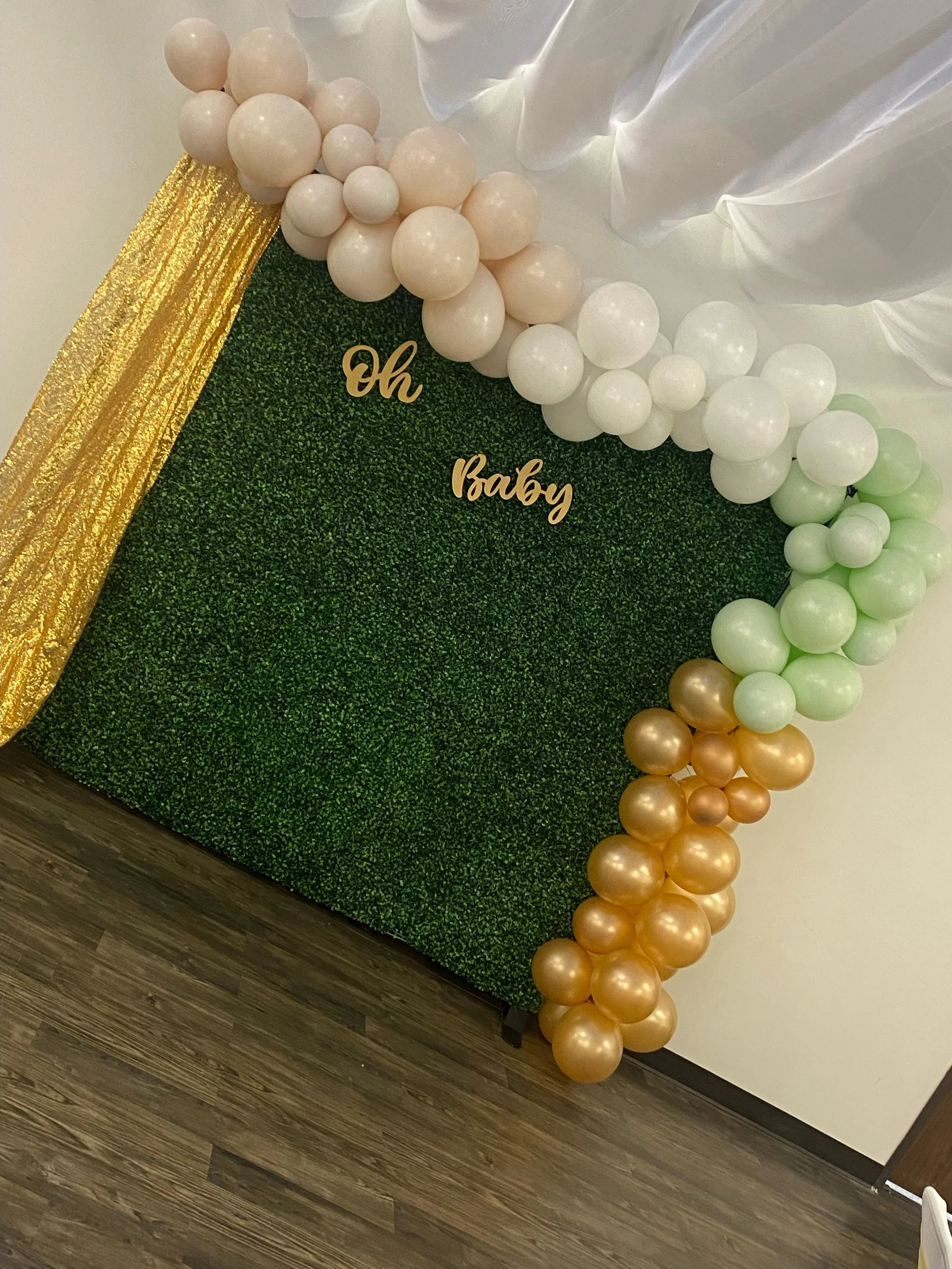 Decorative backdrop with green foliage wall, gold, white, and light green balloons arranged along the edges, and a gold glittery drape. The text 'Oh Baby' is in gold on the foliage wall, indicating a baby shower or similar celebration.
