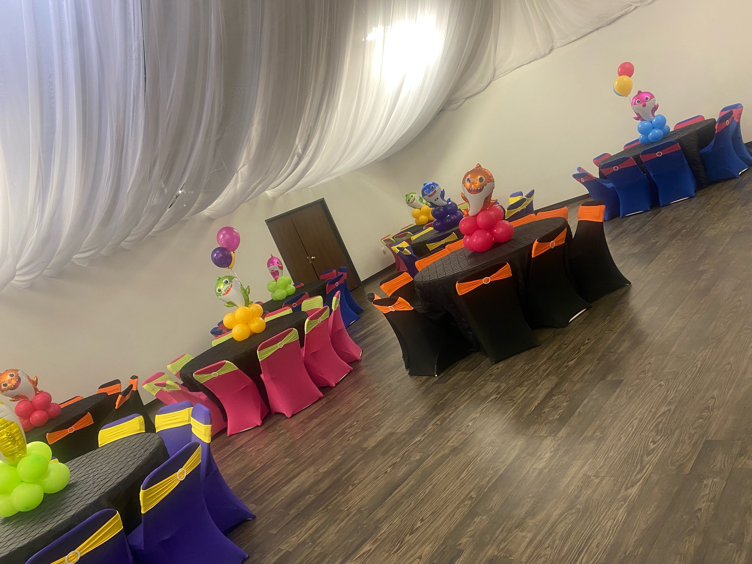 Party room decorated with colorful balloons, balloon sculptures of animals, and decorated chairs with black covers and orange and blue accents.