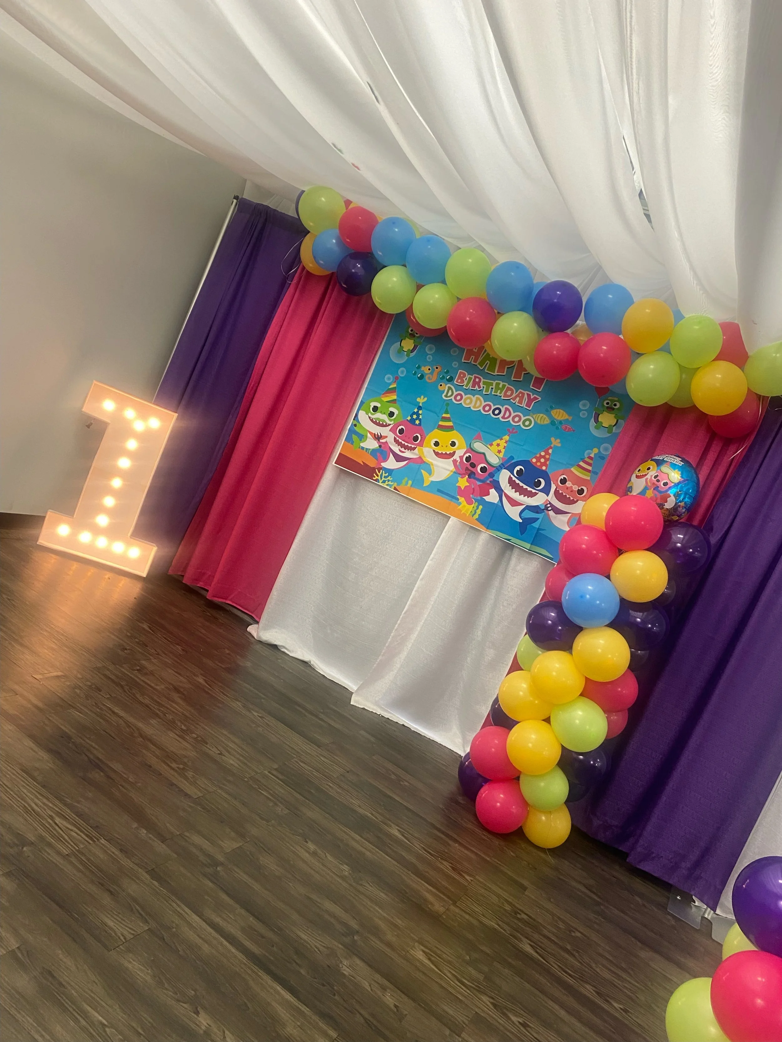 A birthday party decoration with colorful balloons arranged along purple, pink, and white curtains, a large illuminated letter 'L' on the floor, and a cartoon-themed birthday poster with sea creatures and the words 'HAPPY BIRTHDAY DOODOO' in the back