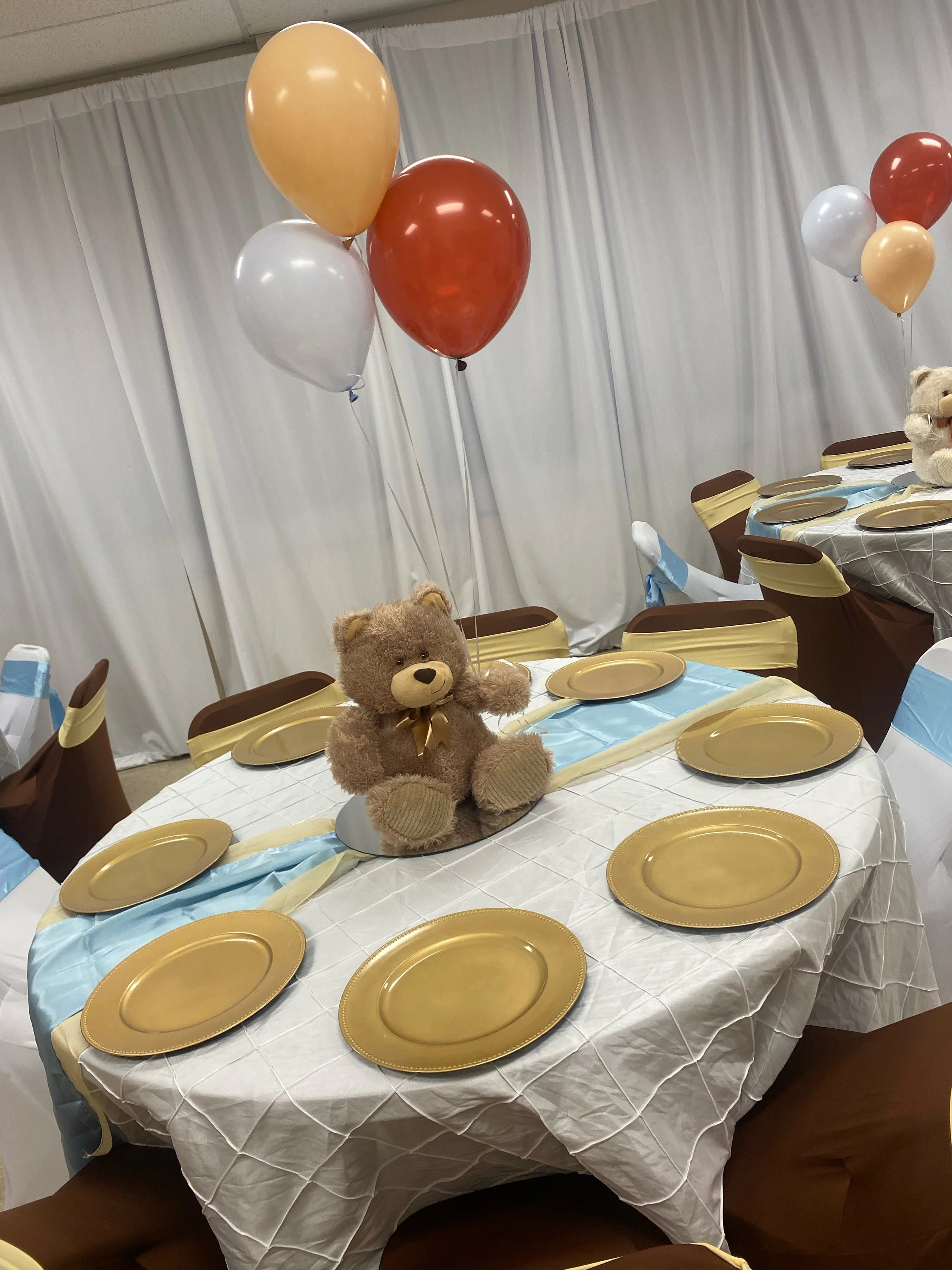 Party table decorated with gold plates, light blue and white tablecloths, teddy bears, balloons in gold, white, and reddish-brown colors.