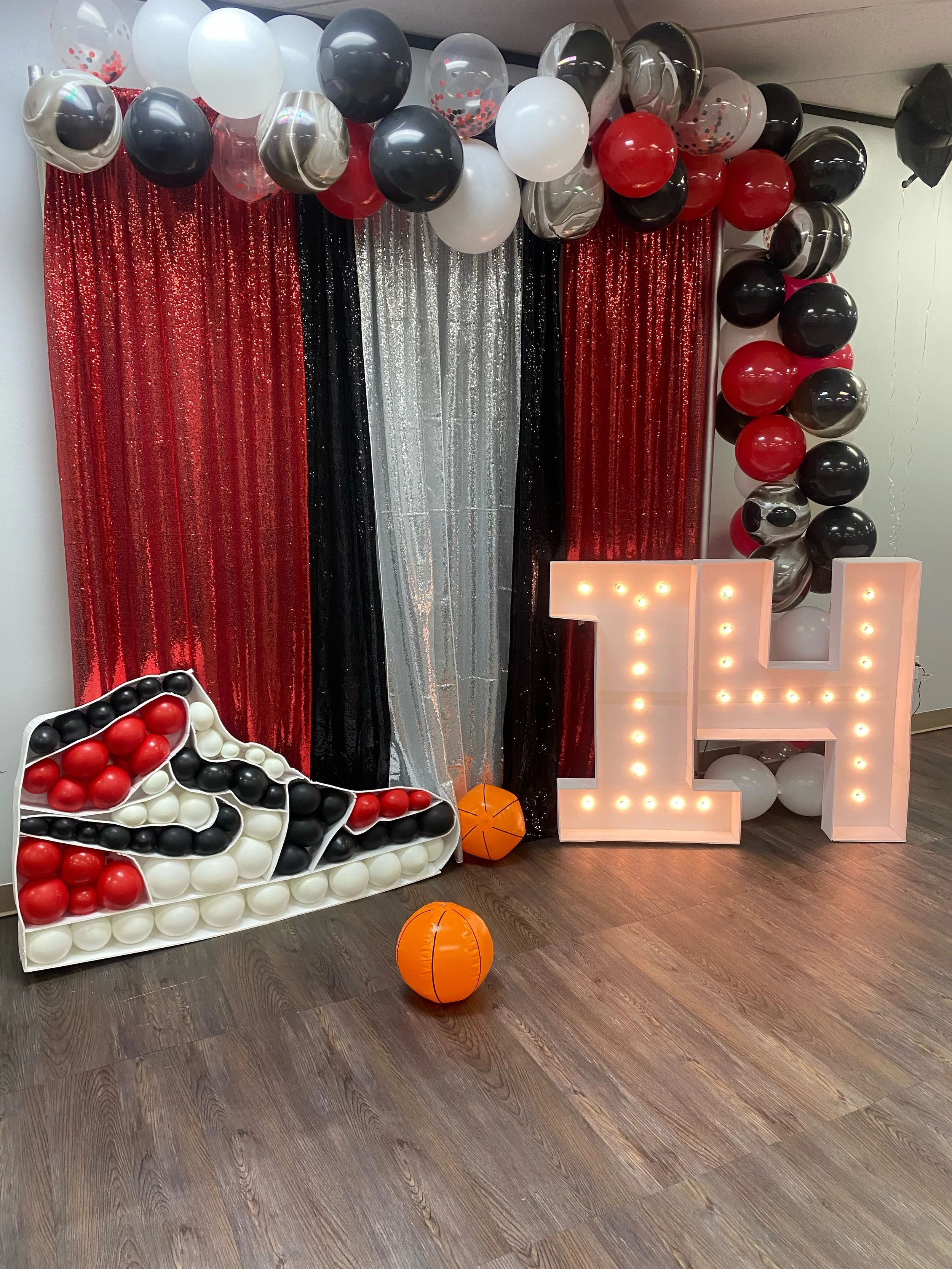 Decorative setup featuring red, white, black, silver, and clear balloons arranged in an arch and around a backdrop of red and black sequin curtains. There is a large pink letter illuminated with bulbs.