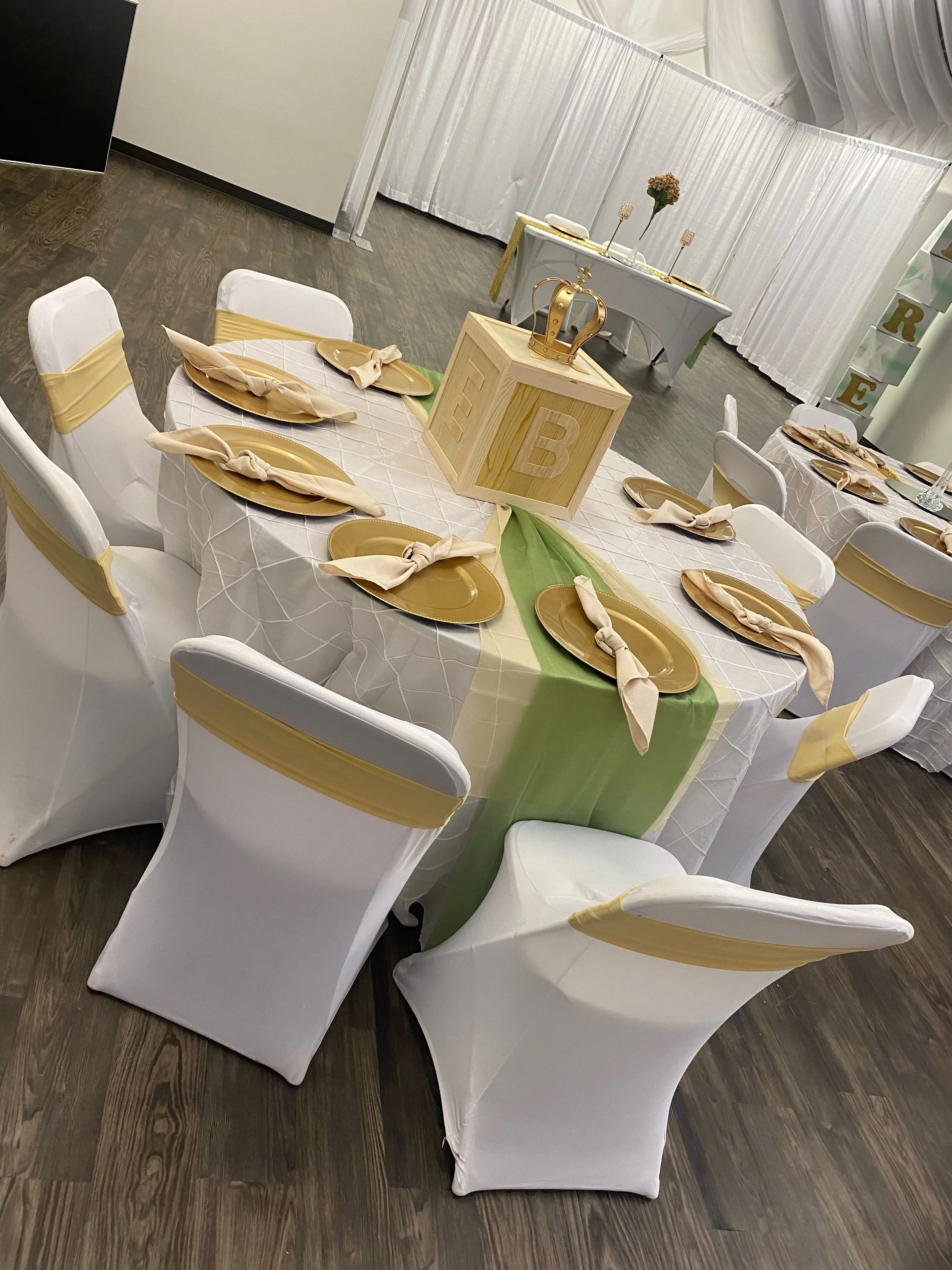 A decorated banquet table with white cloth, gold and beige accents, and a wooden block centerpiece with the letter 'E B' and a gold crown. The table has gold plates and beige napkins with ties. In the background, there's a white draped backdrop with 
