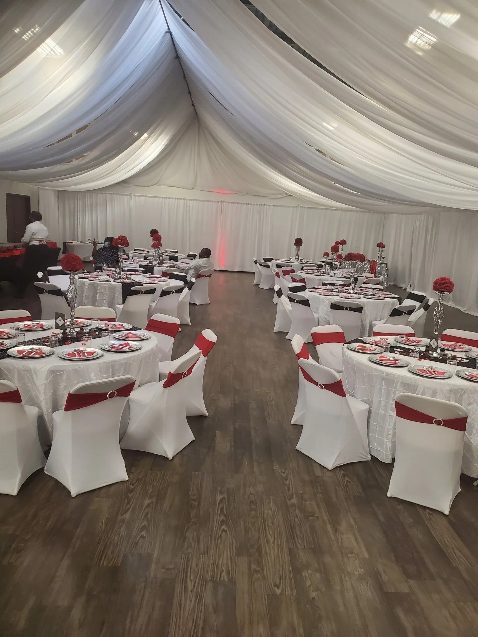 A banquet hall decorated with round tables covered with white tablecloths, each with a tall vase of red flowers. The chairs are white with red sashes, and the ceiling is draped with white fabric. There are a few people preparing the space.