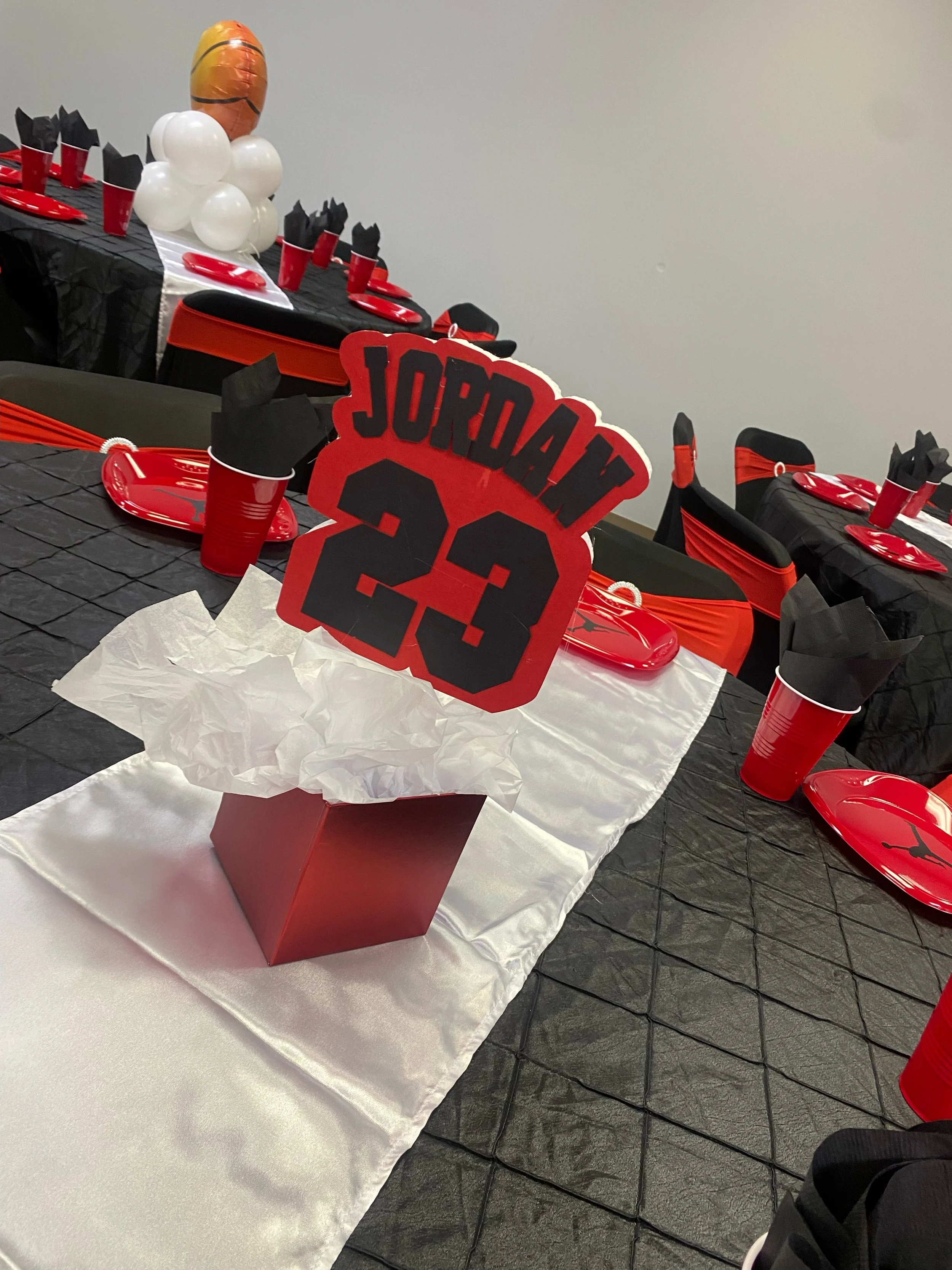 Party table decorated in black and orange with a football theme, featuring balloons, red cups and plates, a cake with a Jordan 23 sign, and black and orange chairs.