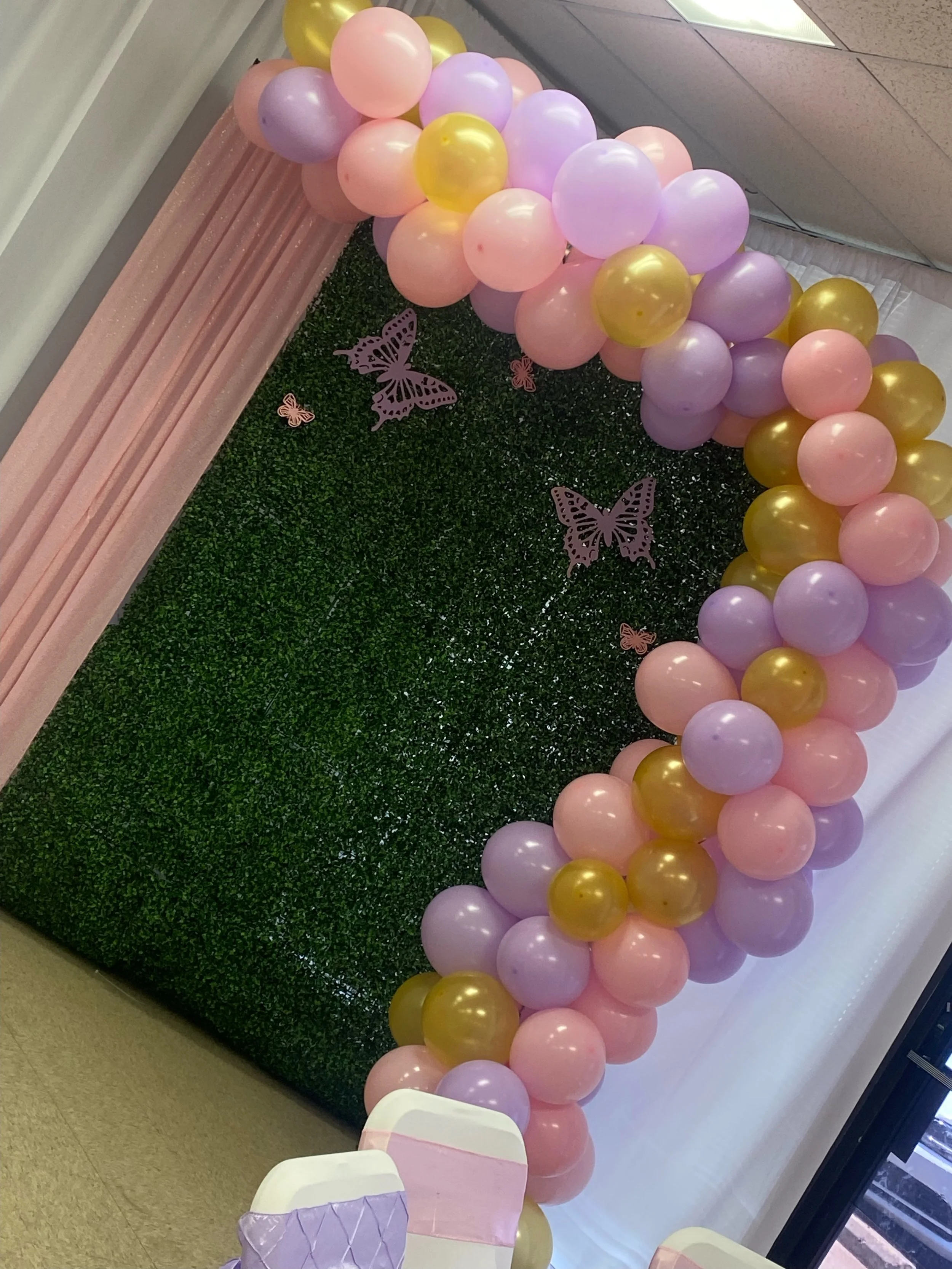 Decorative balloon arch with pink, purple, and yellow balloons, butterflies, and floral accents, set against a green hedge backdrop with pink drapes.