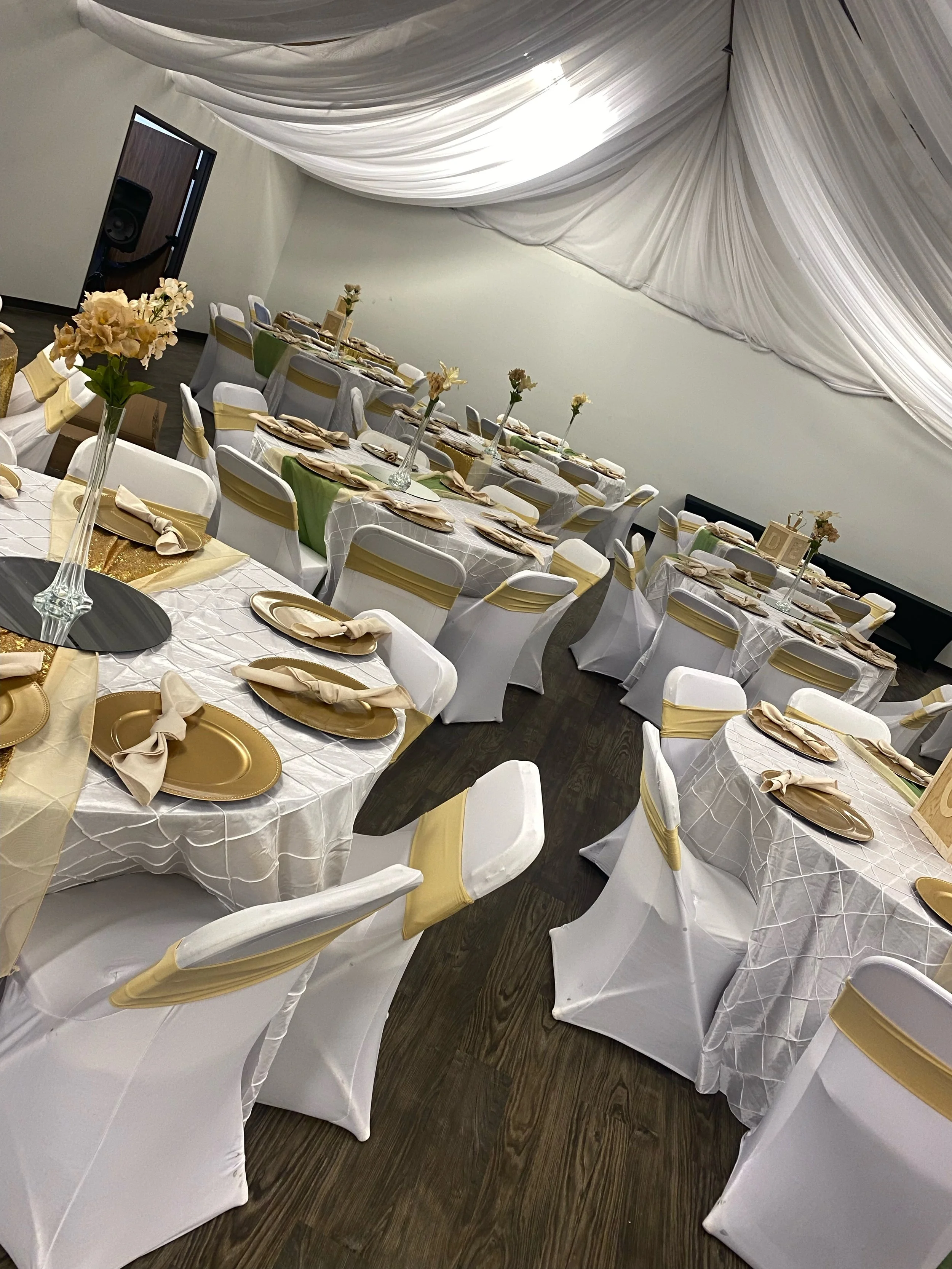 Wedding reception tables decorated with white tablecloths, gold and beige napkins, and centerpieces with beige flowers in tall vases.