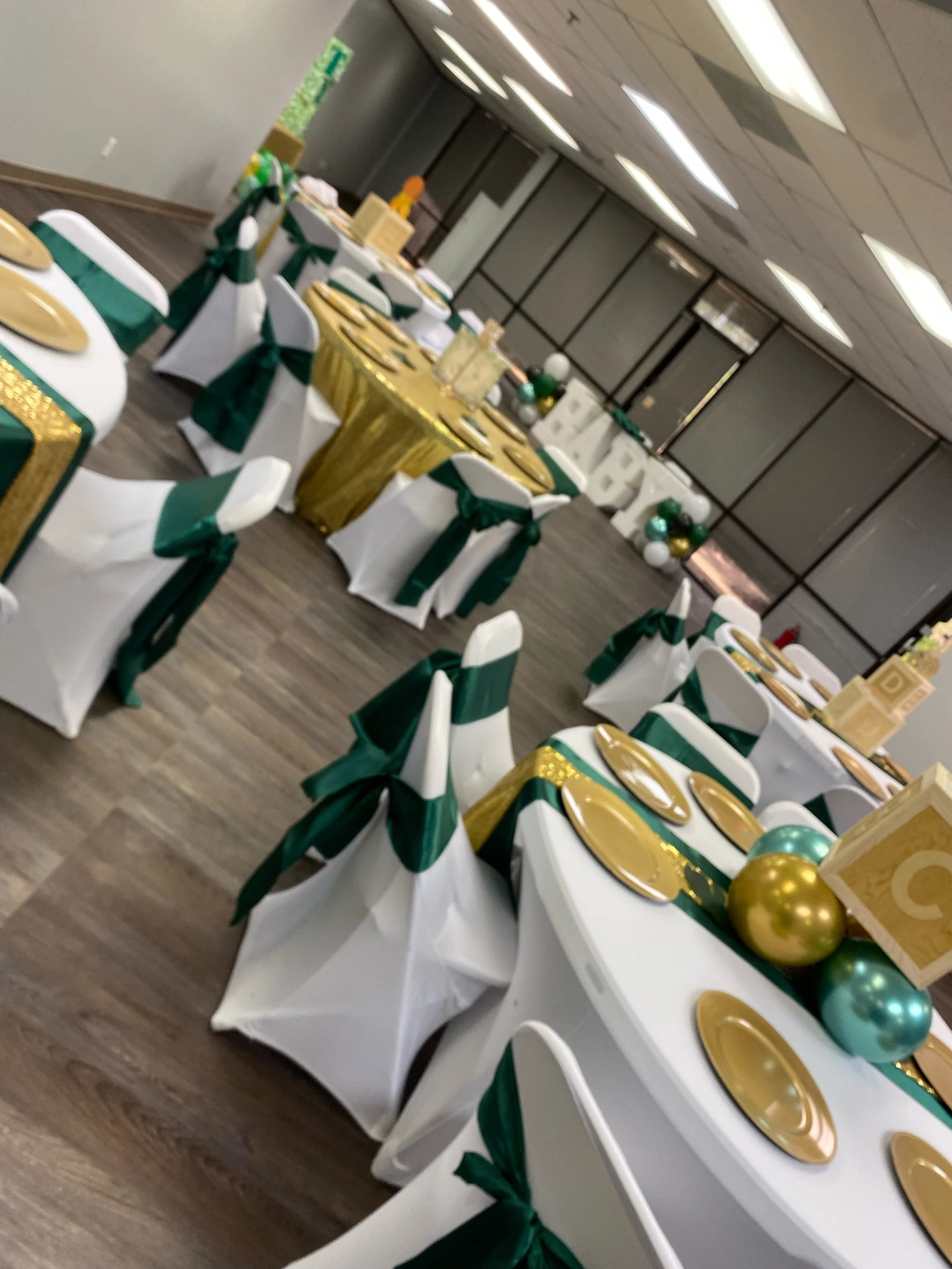 A decorated banquet hall with white chairs adorned with green ribbons, yellow and gold table settings, and balloons in gold, green, and white colors arranged around the tables.