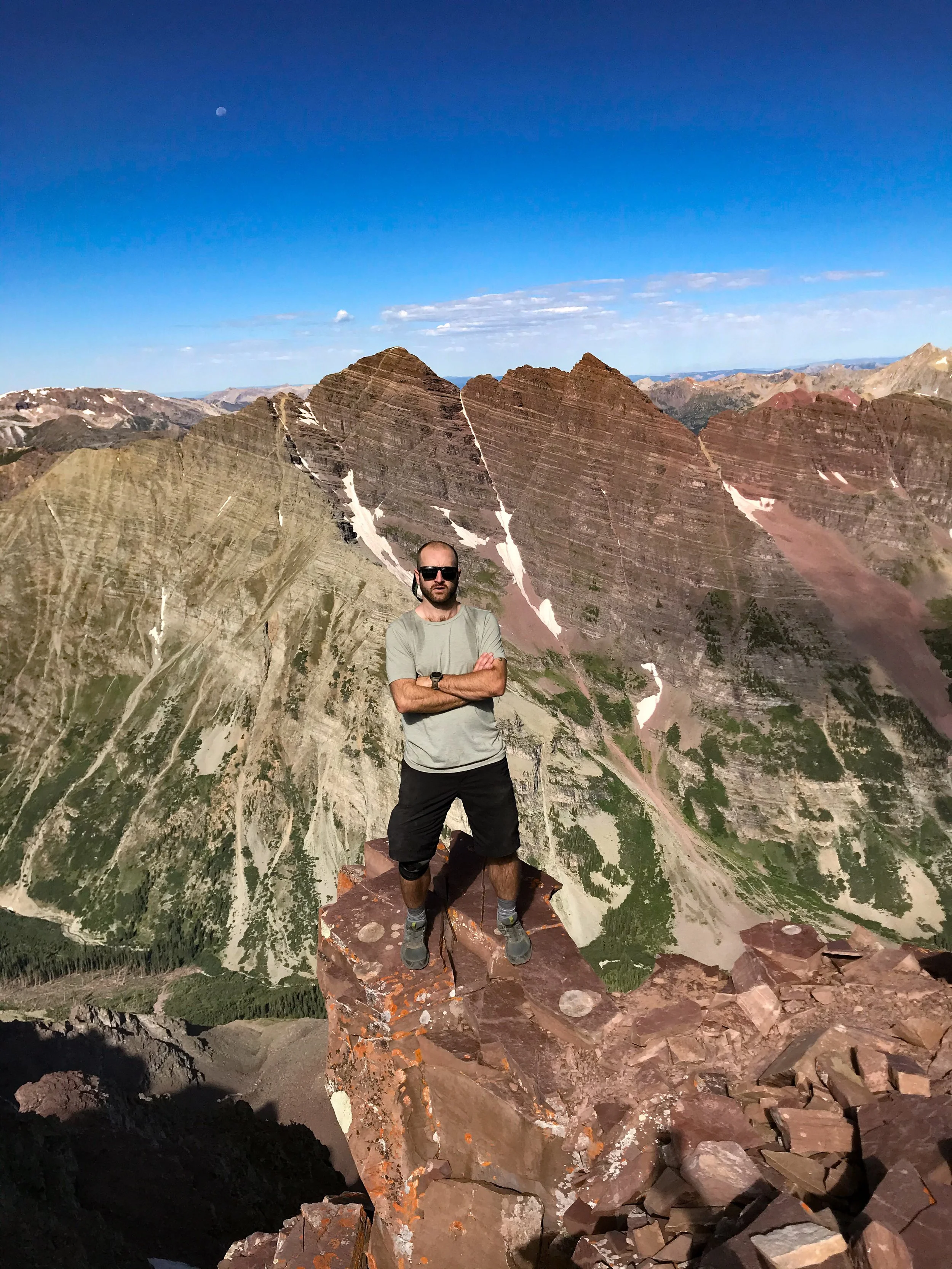 A man standing with arms crossed on a rocky summit, surrounded by mountainous terrain with colorful layered peaks, under a blue sky with a visible moon.