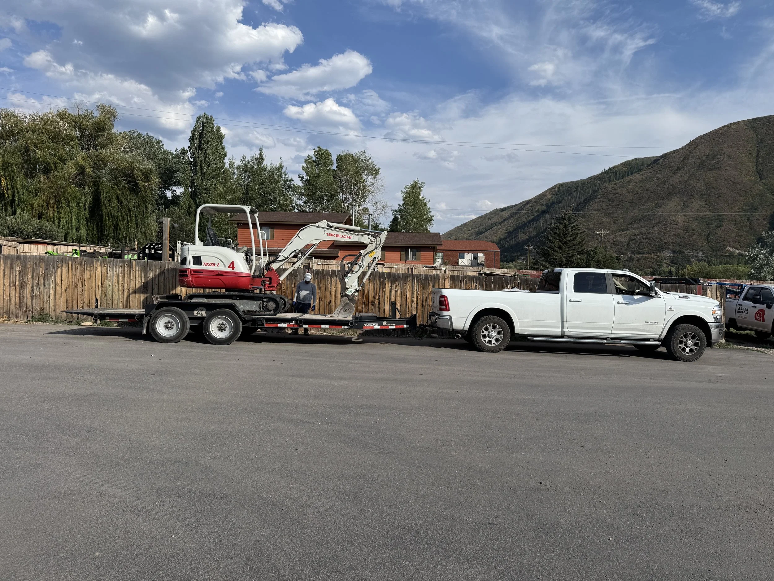 A white pickup truck parked on the side of a road with a trailer carrying a small excavator attached. There is a man walking beside the trailer. In the background, there is a wooden fence, houses, trees, and a mountain under a partly cloudy sky.