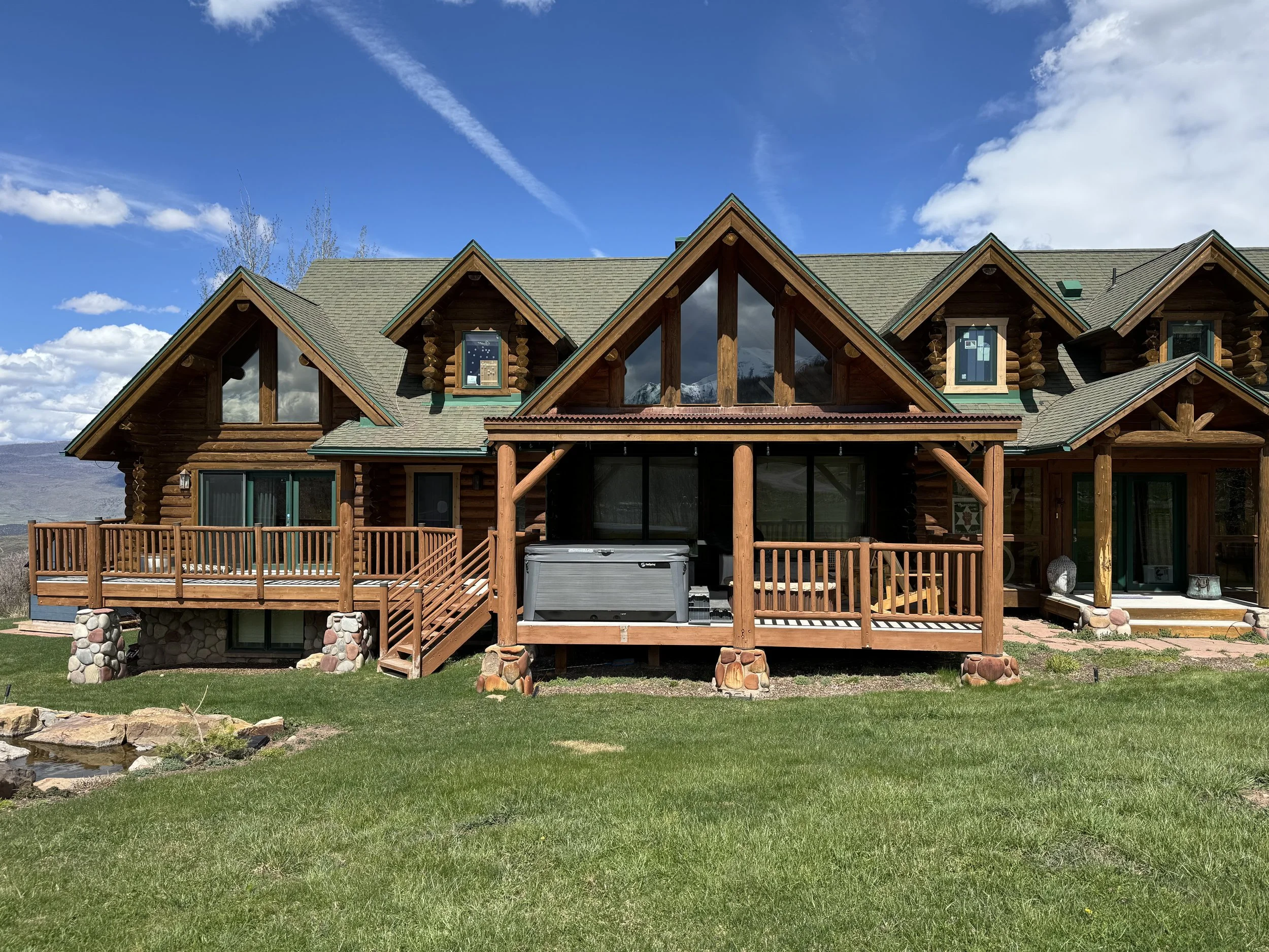 A large wooden house with a deck, multiple gabled roofs, and a hot tub on the deck. The house is set on a grassy lawn with a pond and mountains in the background under a partly cloudy sky.