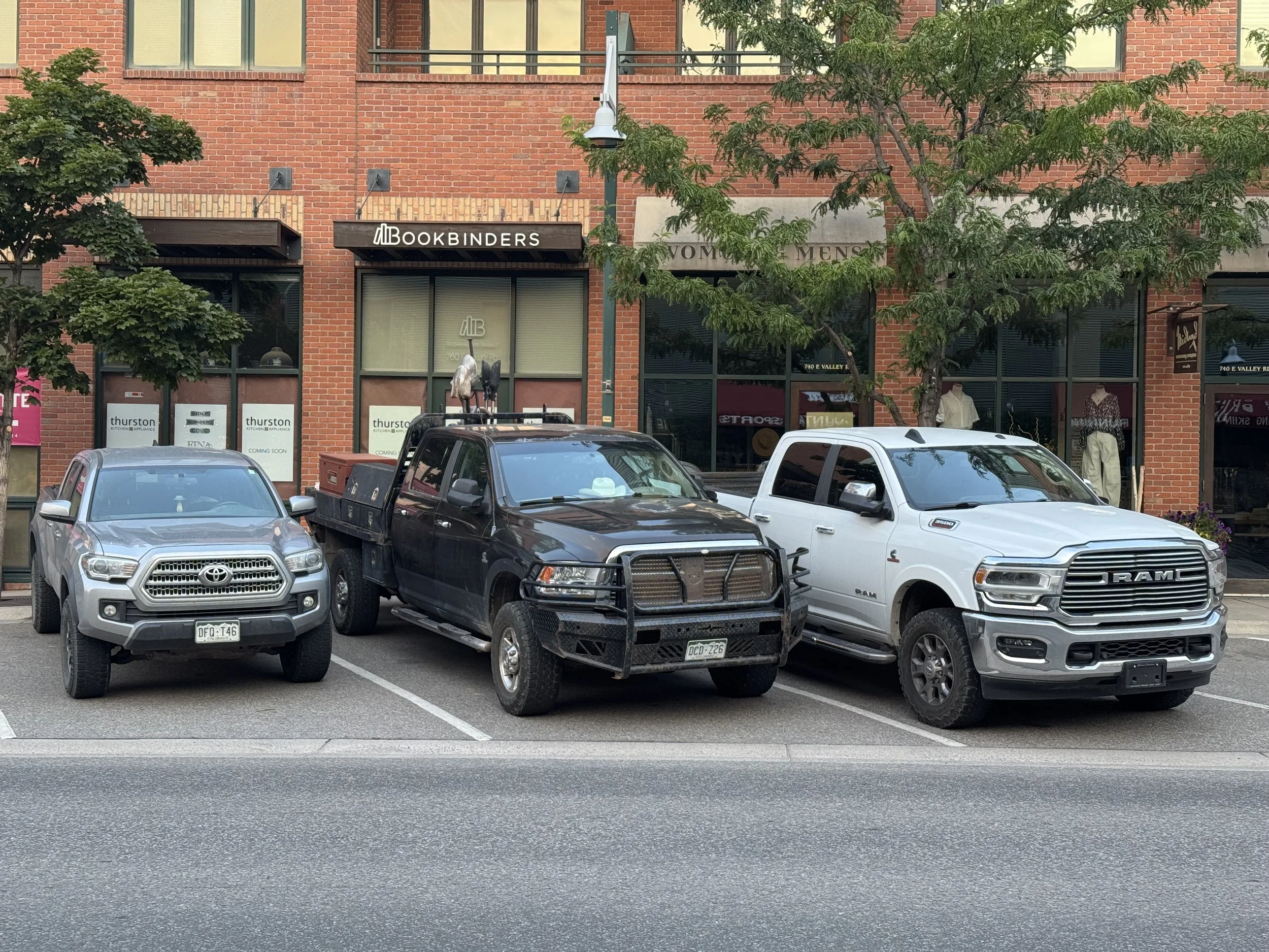 Three trucks parked in front of a brick building with stores and trees. The first truck on the left is silver, the middle one is black with a metal rack, and the right one is white. The building has signs for Bookbinders, Thurston, and women's clothing stores.