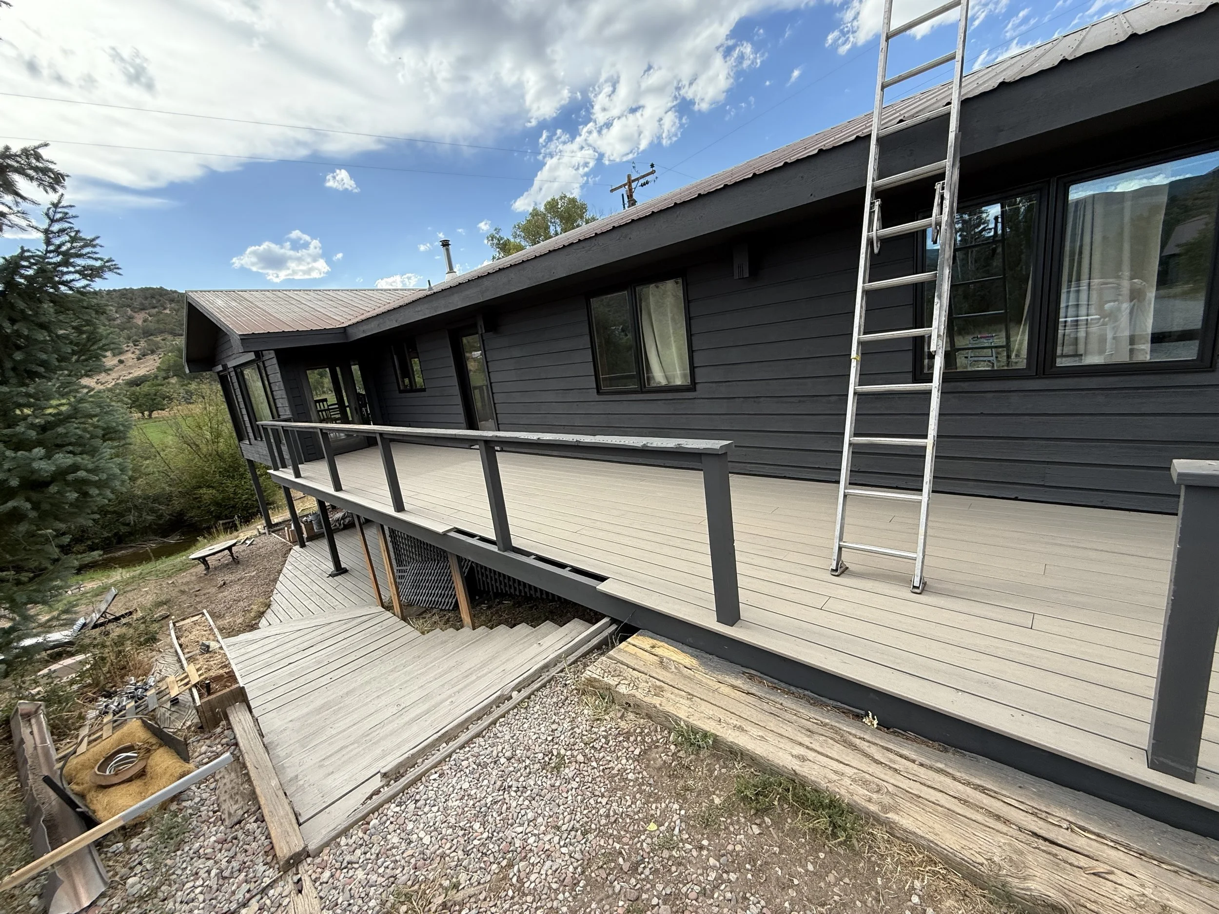 Exterior view of a house with a newly built elevated deck, gray siding, and a ladder leaning against the roof, during daytime with a partly cloudy sky.