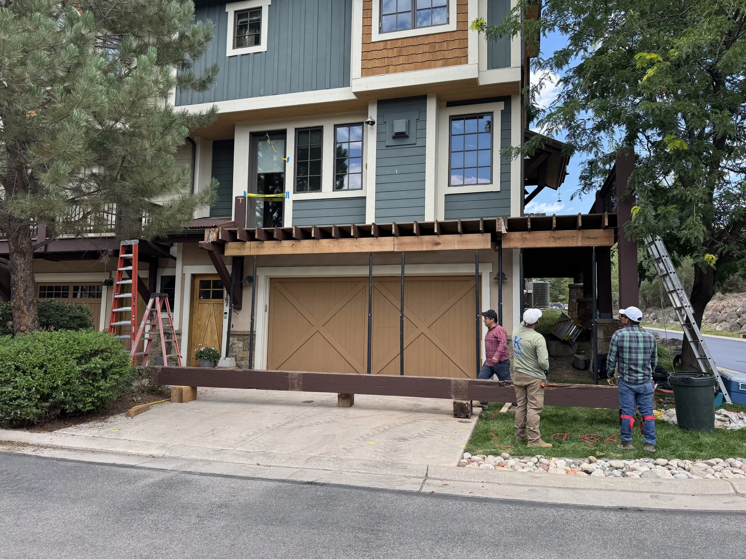 Construction workers standing in front of a house with an unfinished balcony or deck area. There are ladders and construction materials around, and the house features blue and beige exterior siding with large windows.