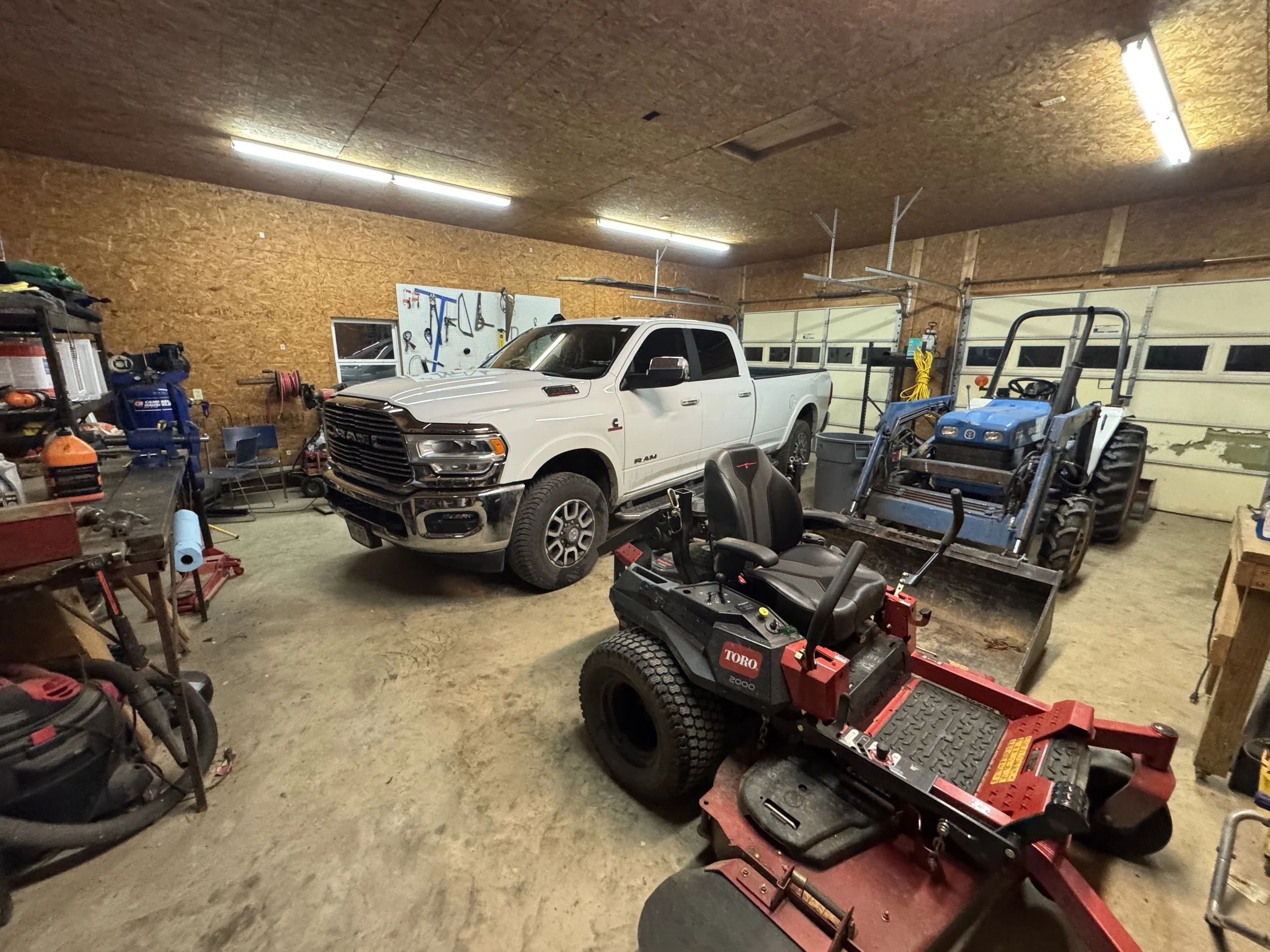 A garage with a white RAM pickup truck, a small blue tractor, and a red Toro riding lawn mower. The garage has shelves, tools, and storage on the walls, with a large garage door at the back.