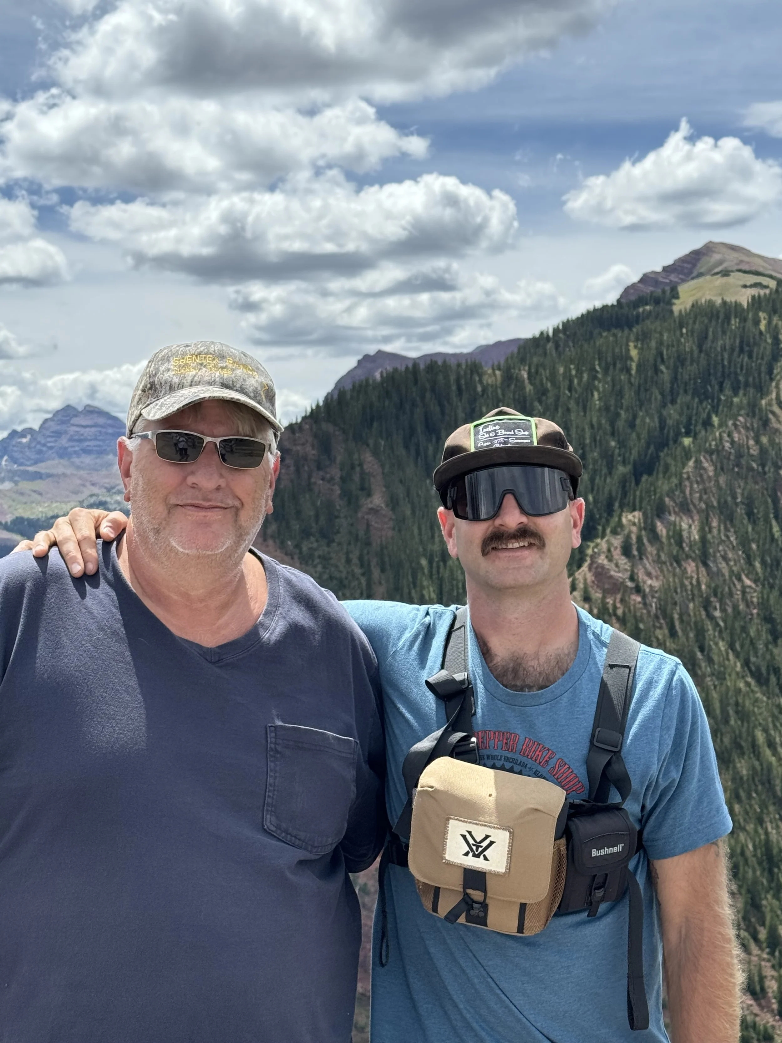 Two men standing together outdoors in a mountainous area with green forested hills and a partly cloudy sky behind them.