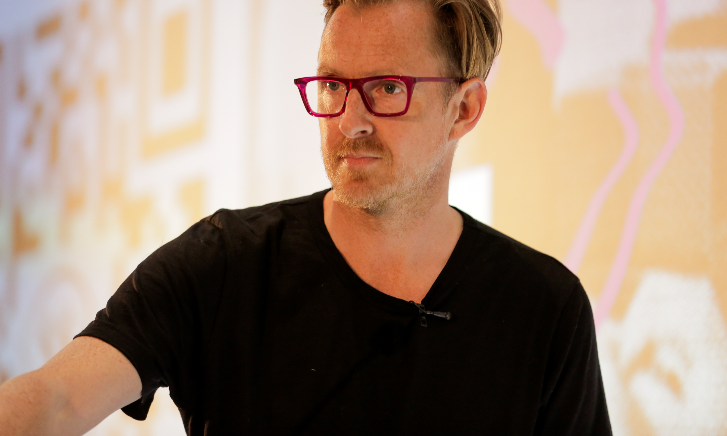 A middle-aged man with red glasses and a black T-shirt standing indoors with a blurred colorful background.