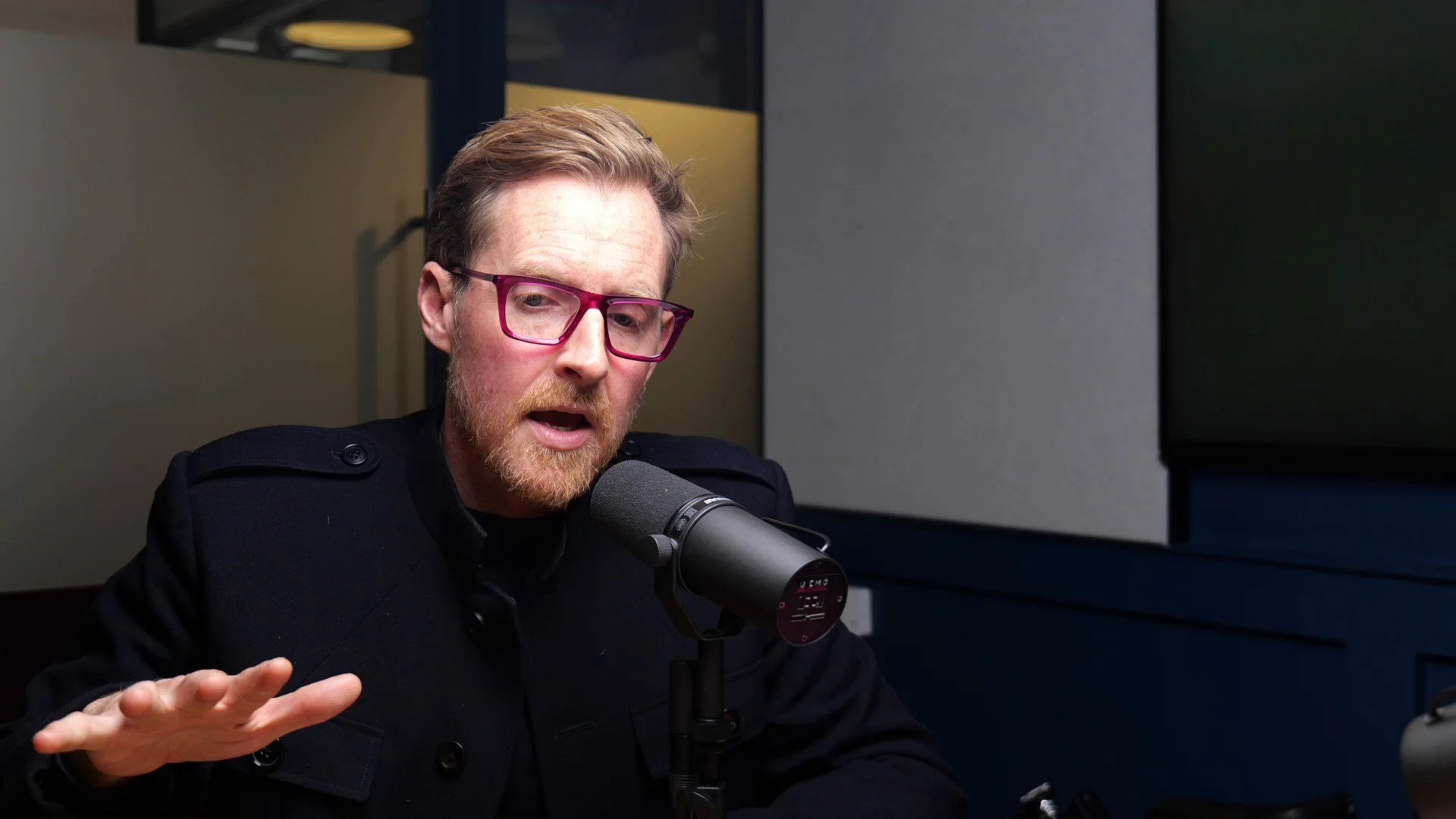 A man with glasses speaking into a microphone in a recording studio.