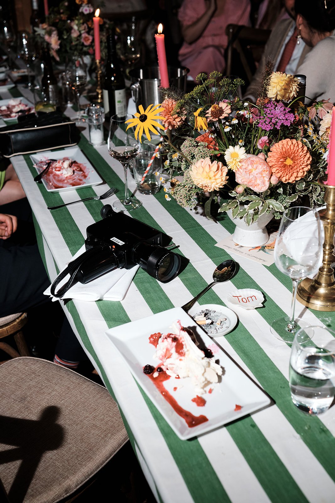 A dining table decorated with a green and white striped tablecloth, featuring a large flower arrangement, pink candles, empty wine glasses, and plates with leftover dessert. A Super 8 camera is placed on the table, and people are sitting around it.