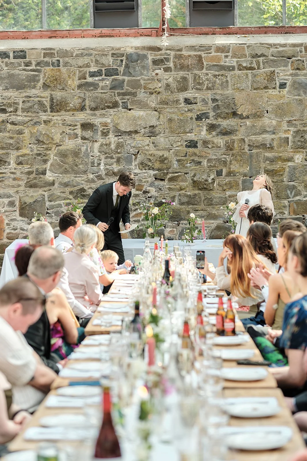 People sitting at a long banquet table, woman in white giving a speech with a microphone, man in black reading from paper, stone wall backdrop, floral decorations, and candles.