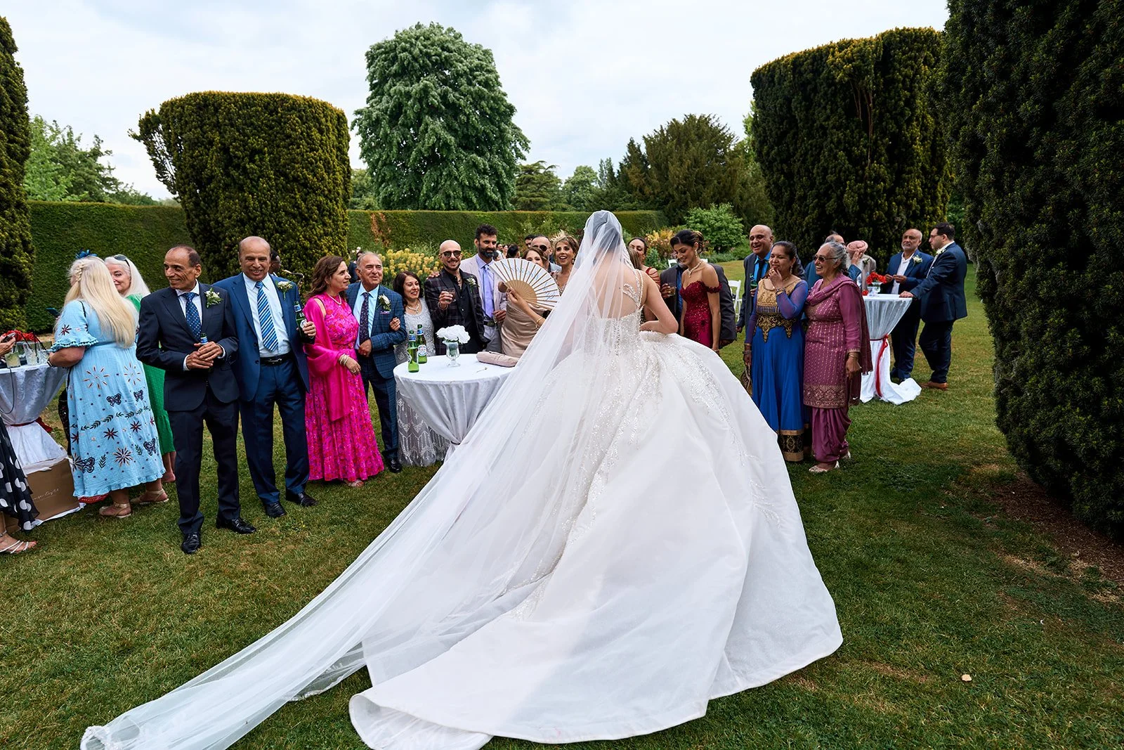 Bride in a white wedding gown with a long veil facing a group of guests outdoors in the east garden at Holme Pierrepont Hall.