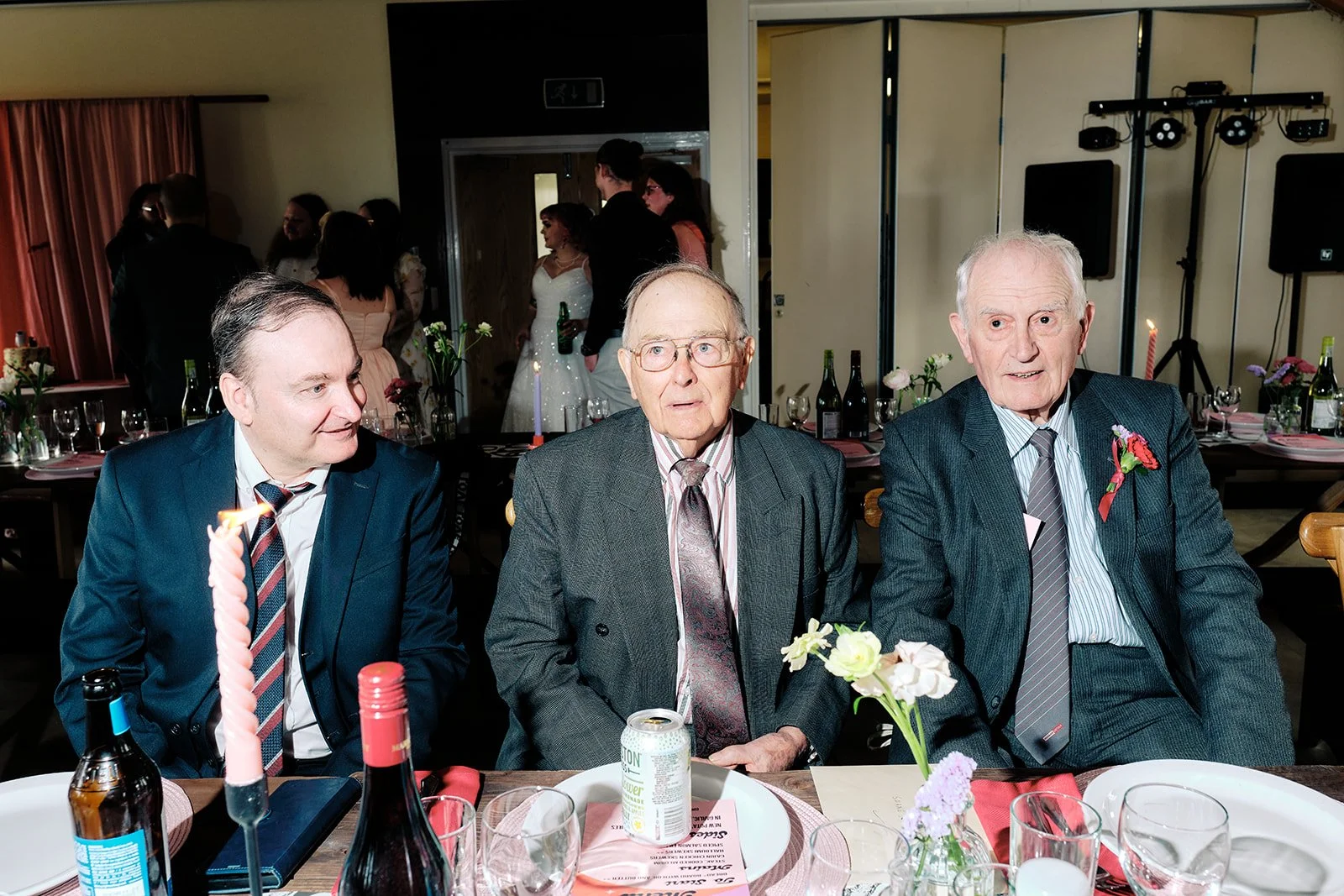 Three elderly men in suits sitting at a decorated dining table during a celebration or wedding reception, with flowers, drinks, and table decor around them.