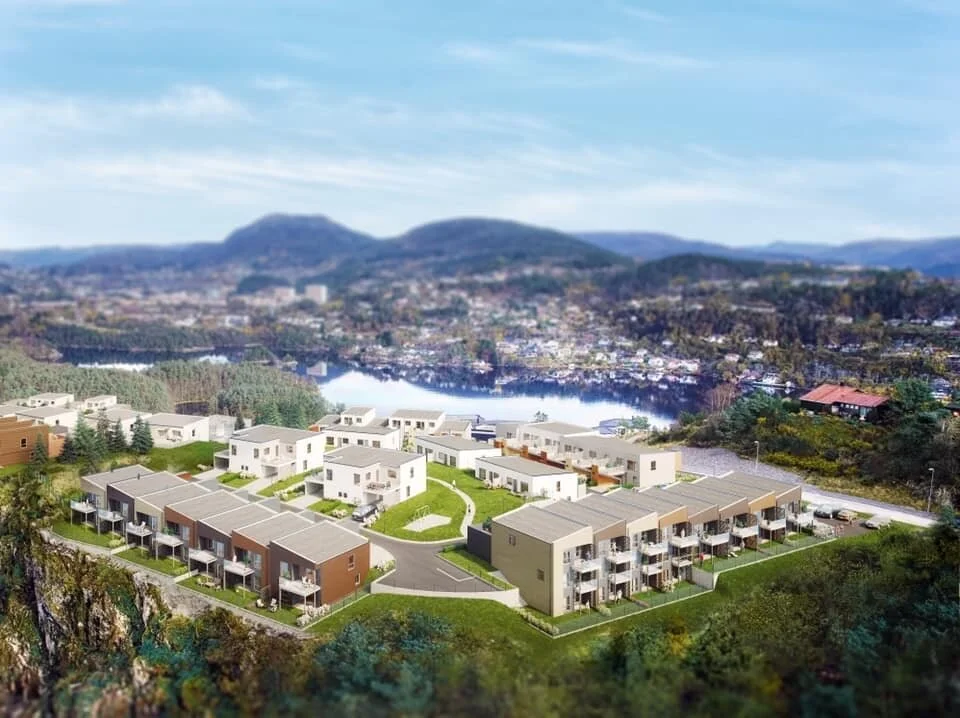 Luftfoto av moderne boligområde med rekkehus og leiligheter, omgitt av natur, innsjø og bakgrunnslandskap.