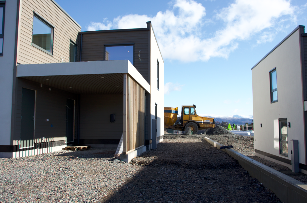 Moderne hus i tre og betong under konstruksjon, med en bulldoser i bakgrunnen. Klart solskinn og fjell i horisonten.