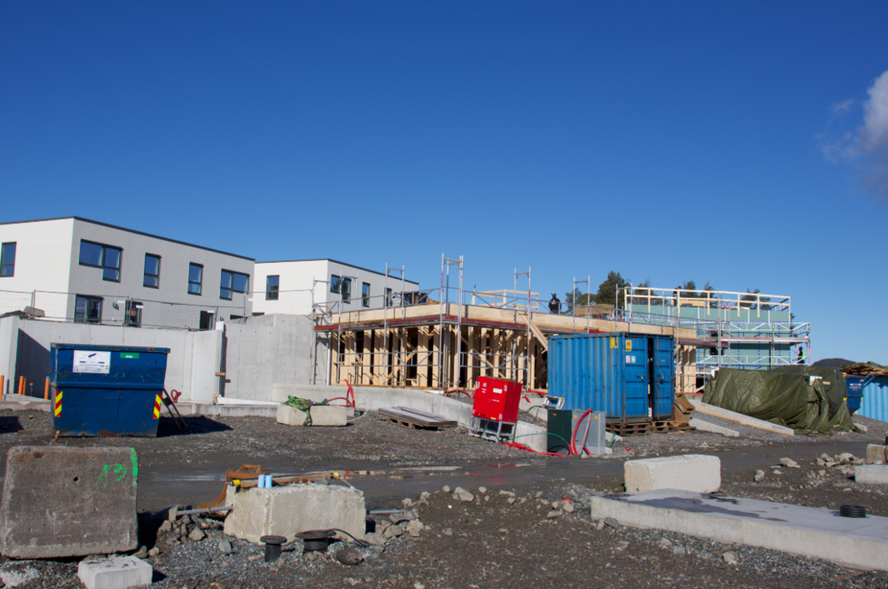 Byggeplass med delvis ferdig bygg, stillas, containere og byggeutstyr. Moderne hvite bygninger i bakgrunnen. Klar blå himmel.