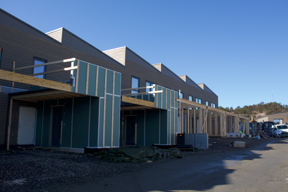 Boligbygging i en rekkehusstruktur under konstruksjon, med trebjelker, takrammer, og isolasjonsplater; byggeplass og blå himmel.
