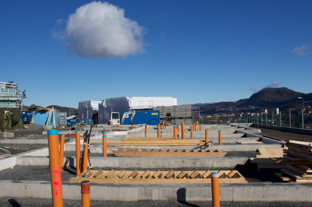 Byggeplass med betongfundamenter, oransje rør, og materialer, med fjell i bakgrunnen.